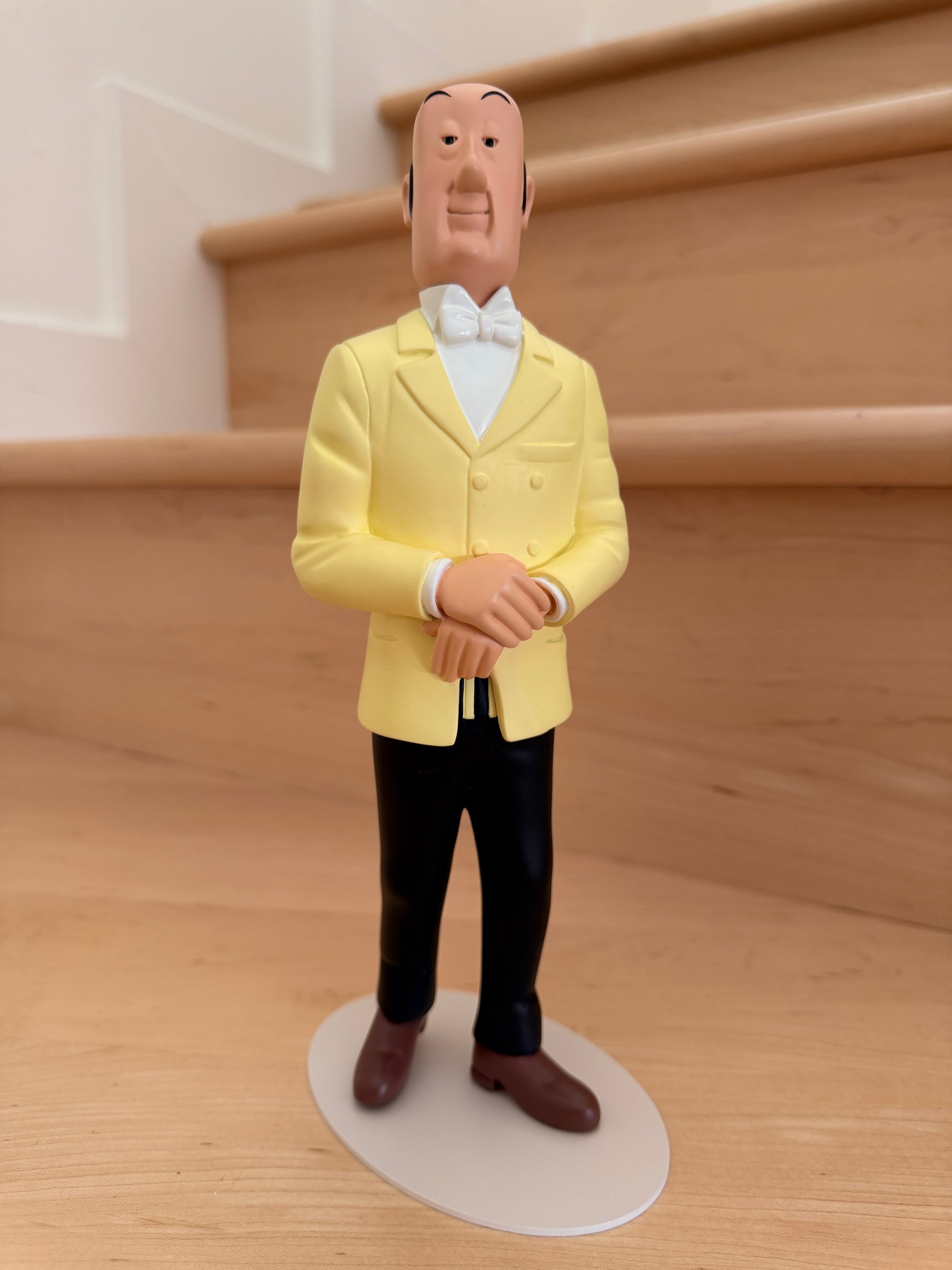 Impeccably dressed Tintinimaginatio Icons collection resin figurine of Nestor, the butler of Marlinspike Hall from The Adventures of Tintin. He is wearing his classic yellow double-breasted suit jacket, white shirt, and white bowtie, standing poised with his hands clasped calmly in front of him on a minimalist circular ba