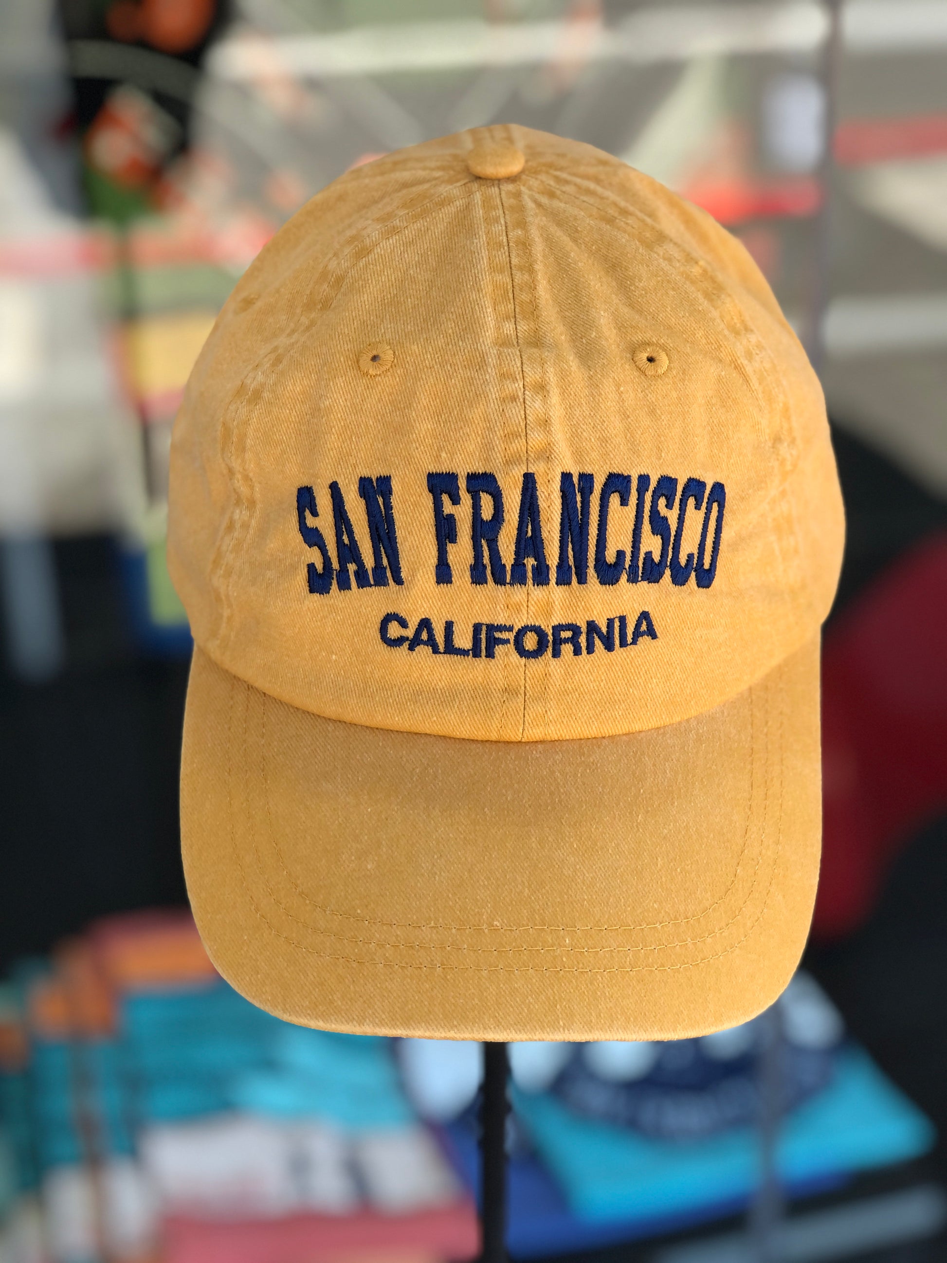Front-facing view of a mango yellow washed cotton baseball cap featuring the words 'San Francisco' in a bold navy blue arched font with 'California' embroidered directly underneath.