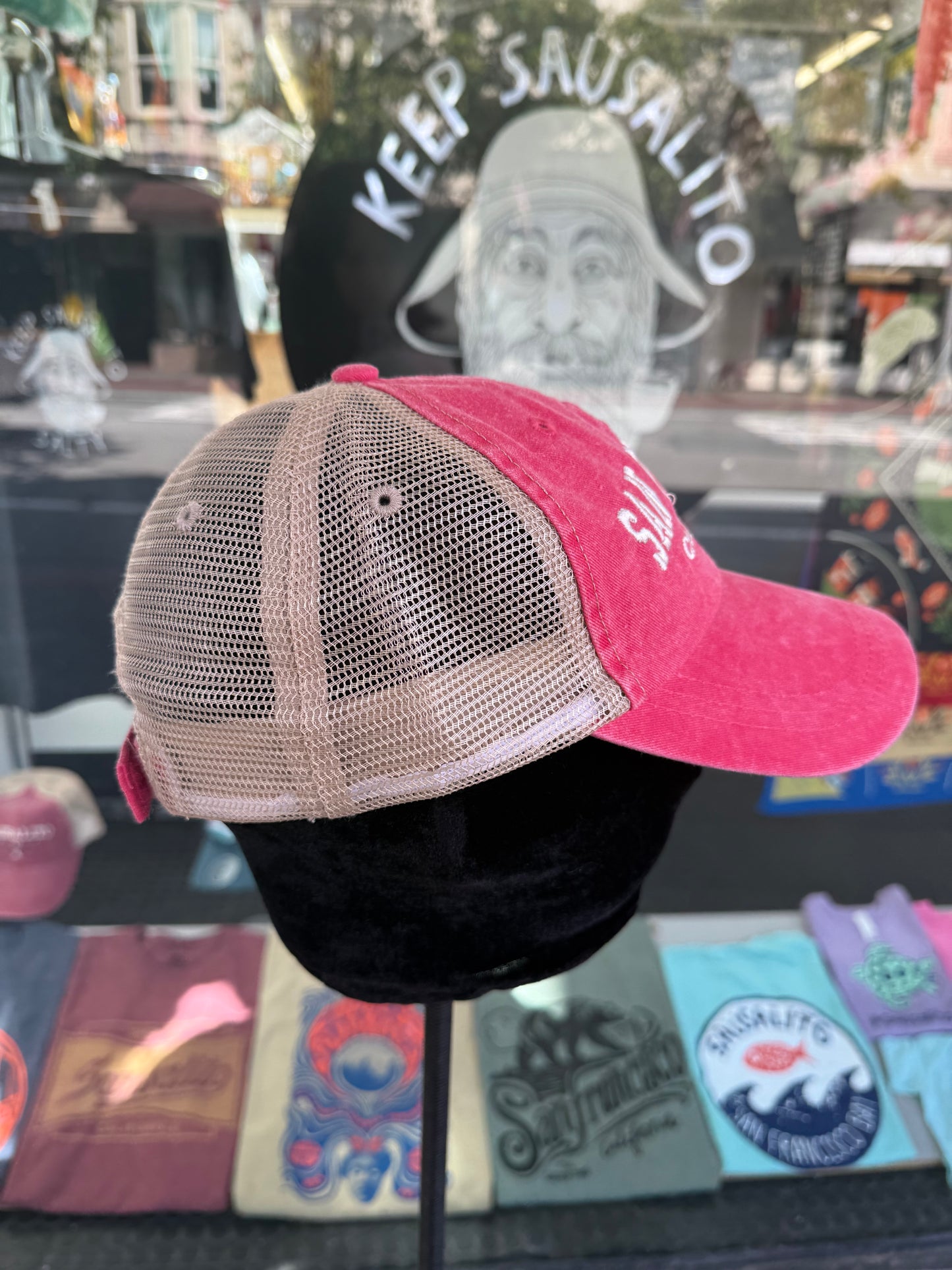 Side profile view of a pink over-washed trucker hat displayed on a mannequin head inside a store window. The hat features a pink brim, a pink front panel with white embroidered lettering, and light beige mesh back panels.