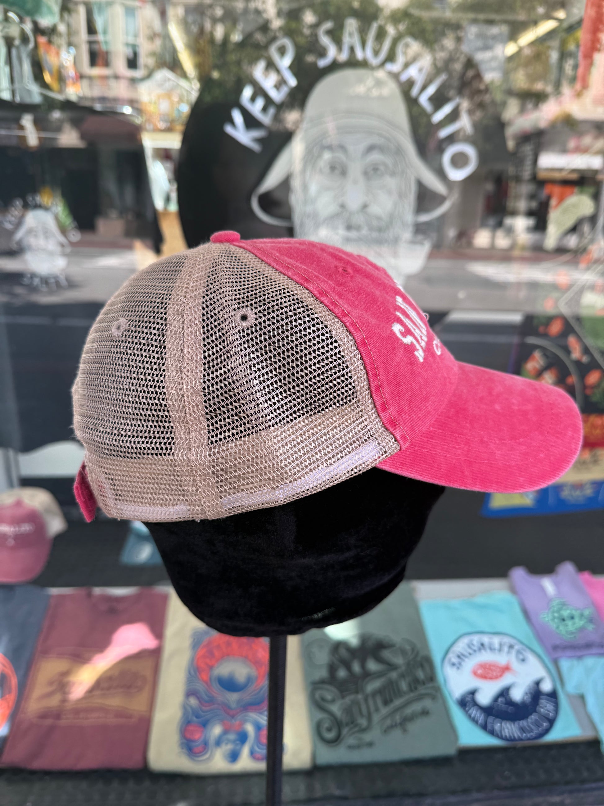 Side profile view of a pink over-washed trucker hat displayed on a mannequin head inside a store window. The hat features a pink brim, a pink front panel with white embroidered lettering, and light beige mesh back panels.