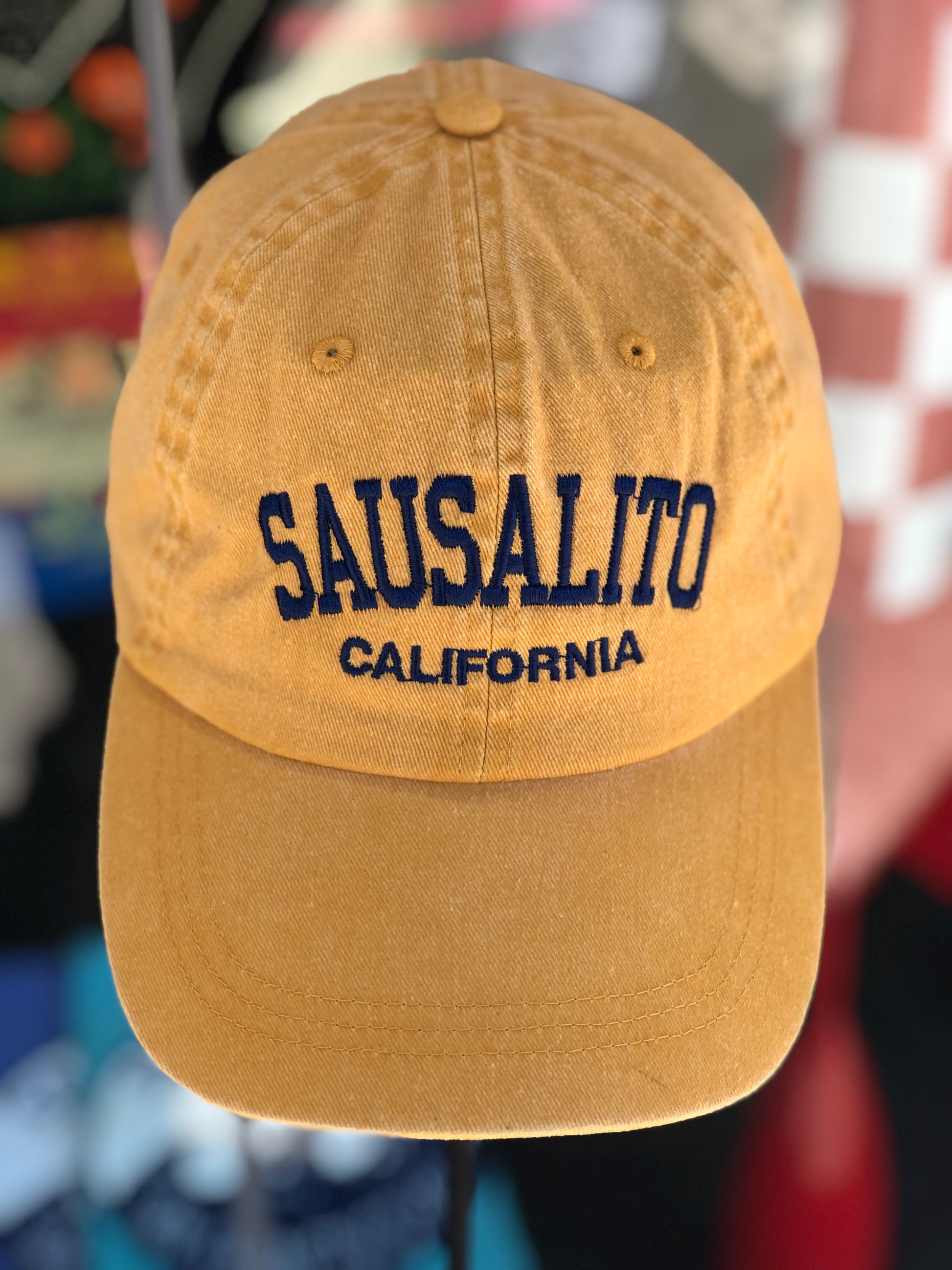 A close-up view of a mango yellow over-washed cotton baseball cap featuring the words 'Sausalito' in a bold navy blue arched font with 'California' embroidered directly underneath.