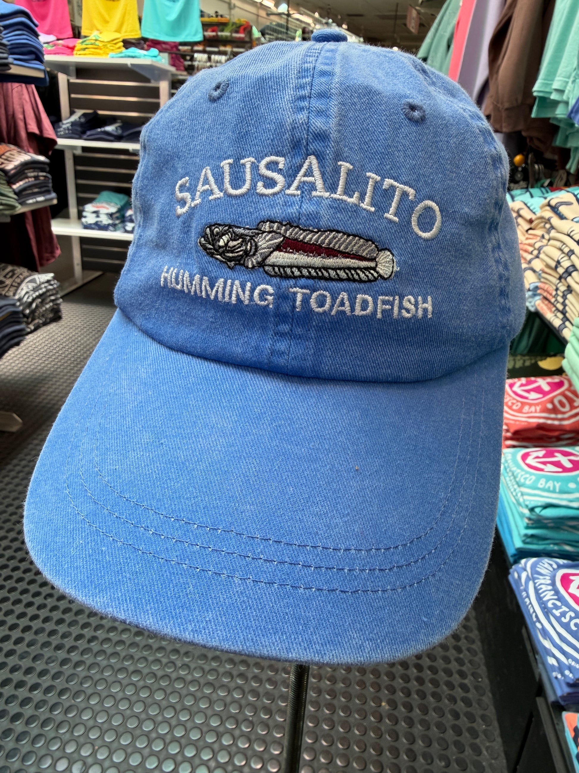 Close-up of a washed royal blue trucker cap featuring an embroidered Sausalito Humming Toadfish logo on a store display shelf.