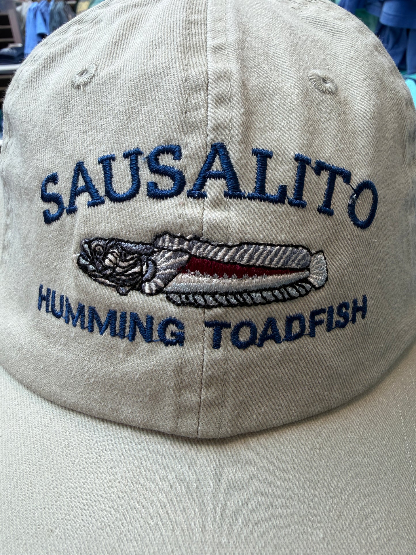Close-up of a beige over-washed cotton trucker hat featuring navy blue embroidered text reading Sausalito Humming Toadfish around a detailed toadfish illustration.