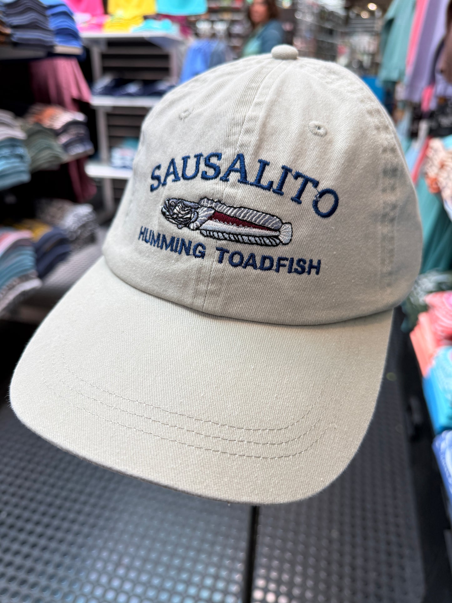 Close-up of a beige over-washed cotton and mesh trucker hat featuring white embroidered text reading Sausalito Humming Toadfish around a detailed toadfish illustration.