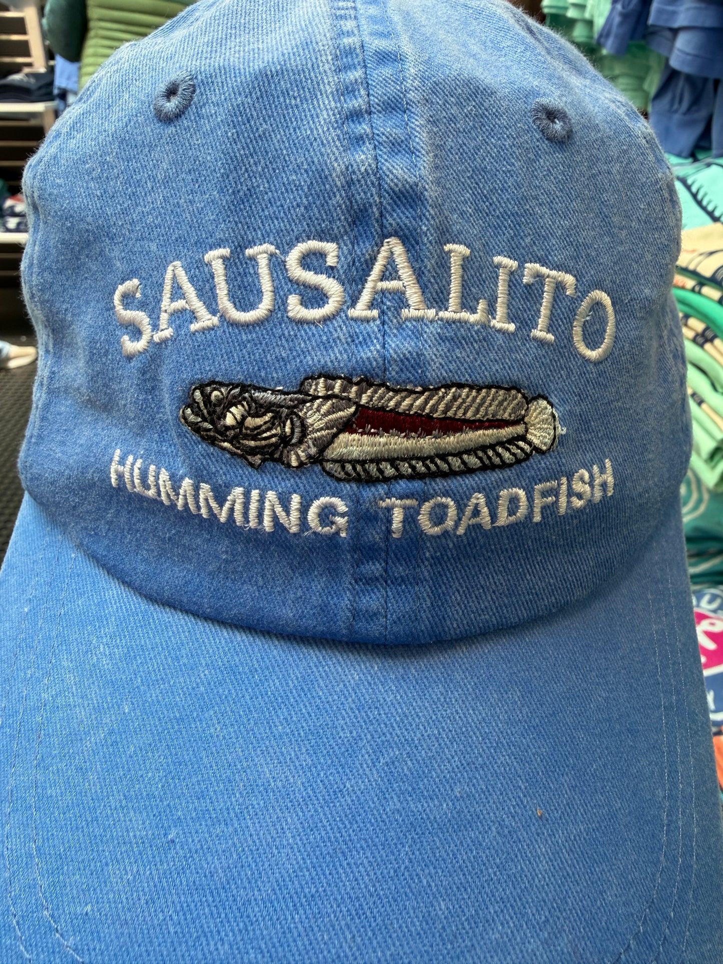 Close-up of a blue over-washed cotton and mesh trucker hat featuring white embroidered text reading Sausalito Humming Toadfish around a detailed toadfish illustration.
