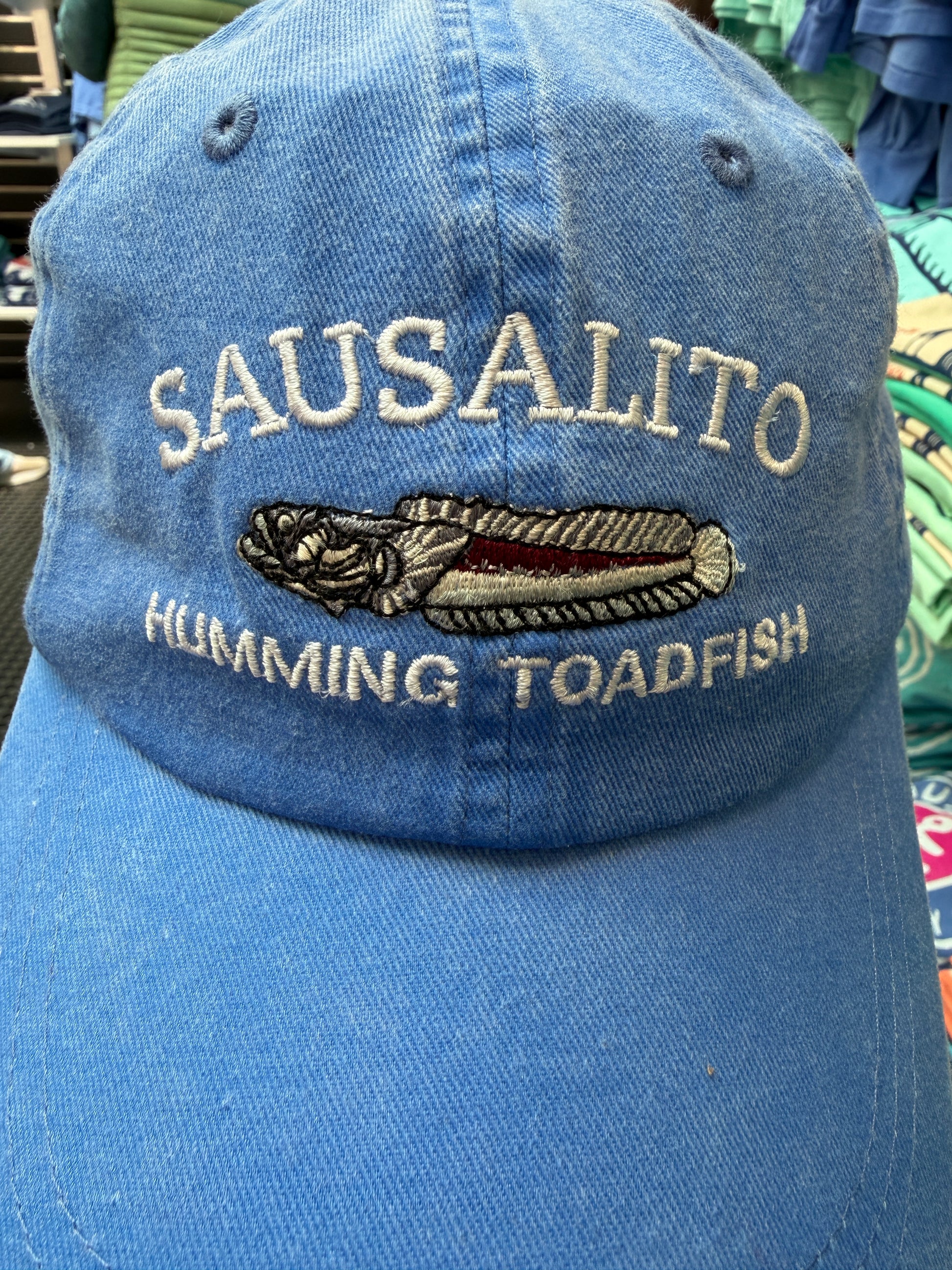 Close-up of a blue over-washed cotton and mesh trucker hat featuring white embroidered text reading Sausalito Humming Toadfish around a detailed toadfish illustration.