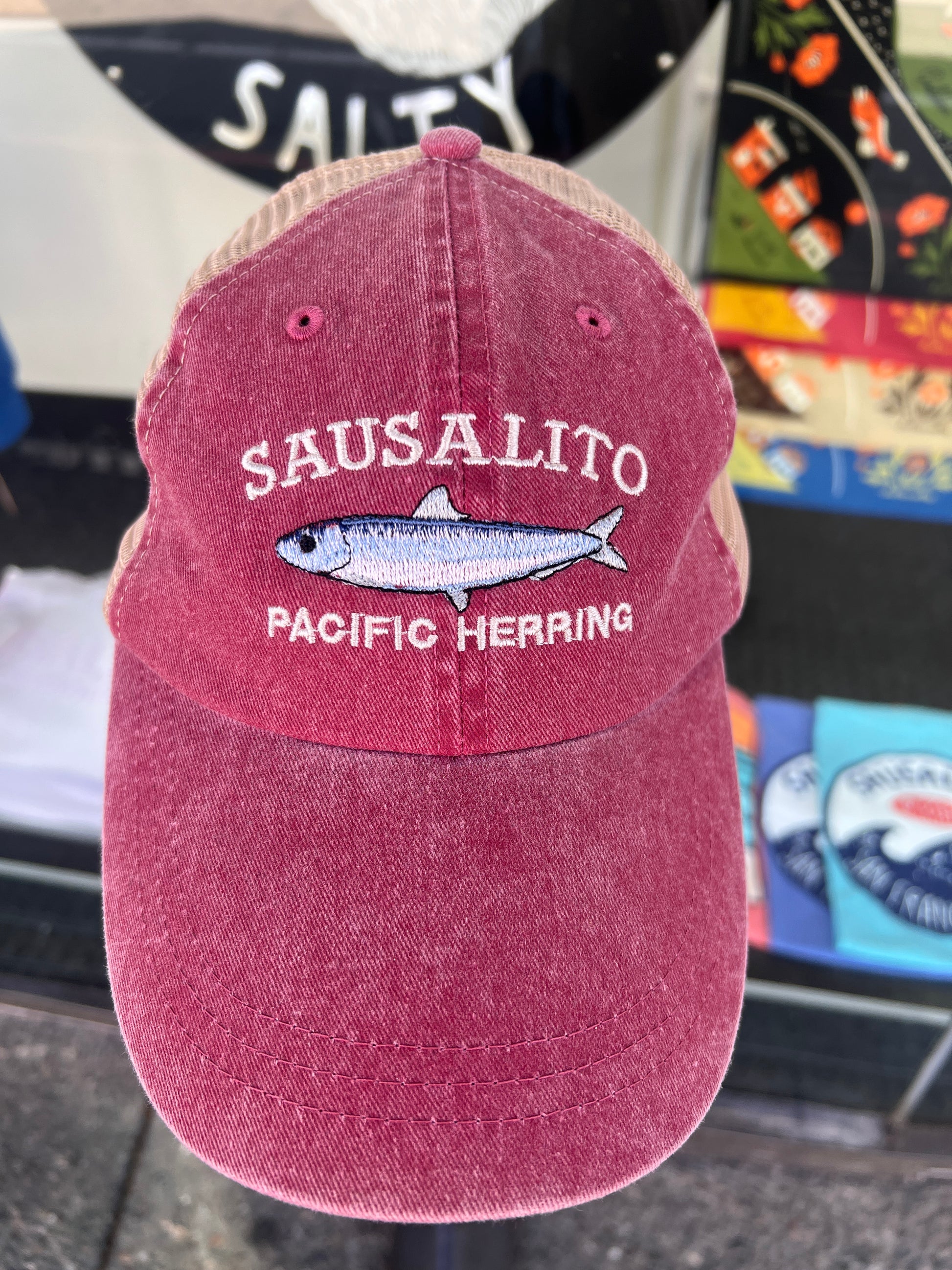 Brick red washed cotton trucker hat featuring an embroidered white "SAUSALITO PACIFIC HERRING" design and a detailed silver-and-blue fish graphic.