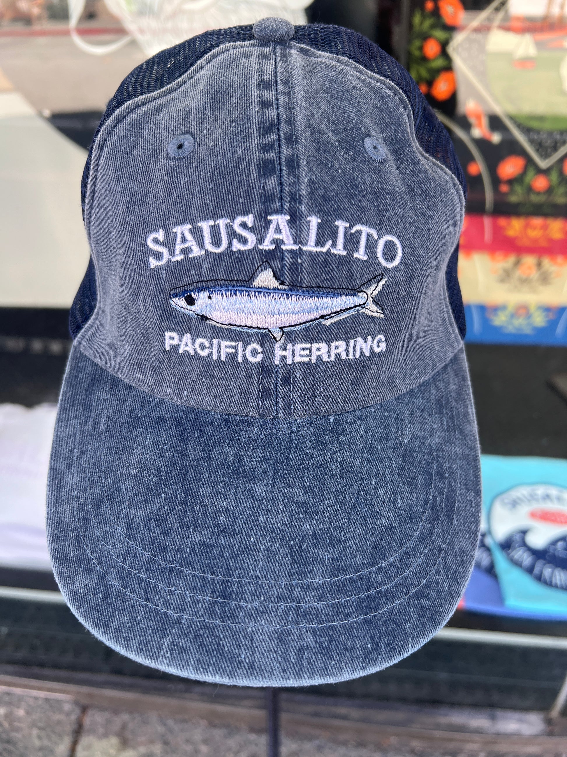 Blue washed denim trucker hat featuring an embroidered white "SAUSALITO PACIFIC HERRING" design with a detailed silver and blue herring fish graphic.