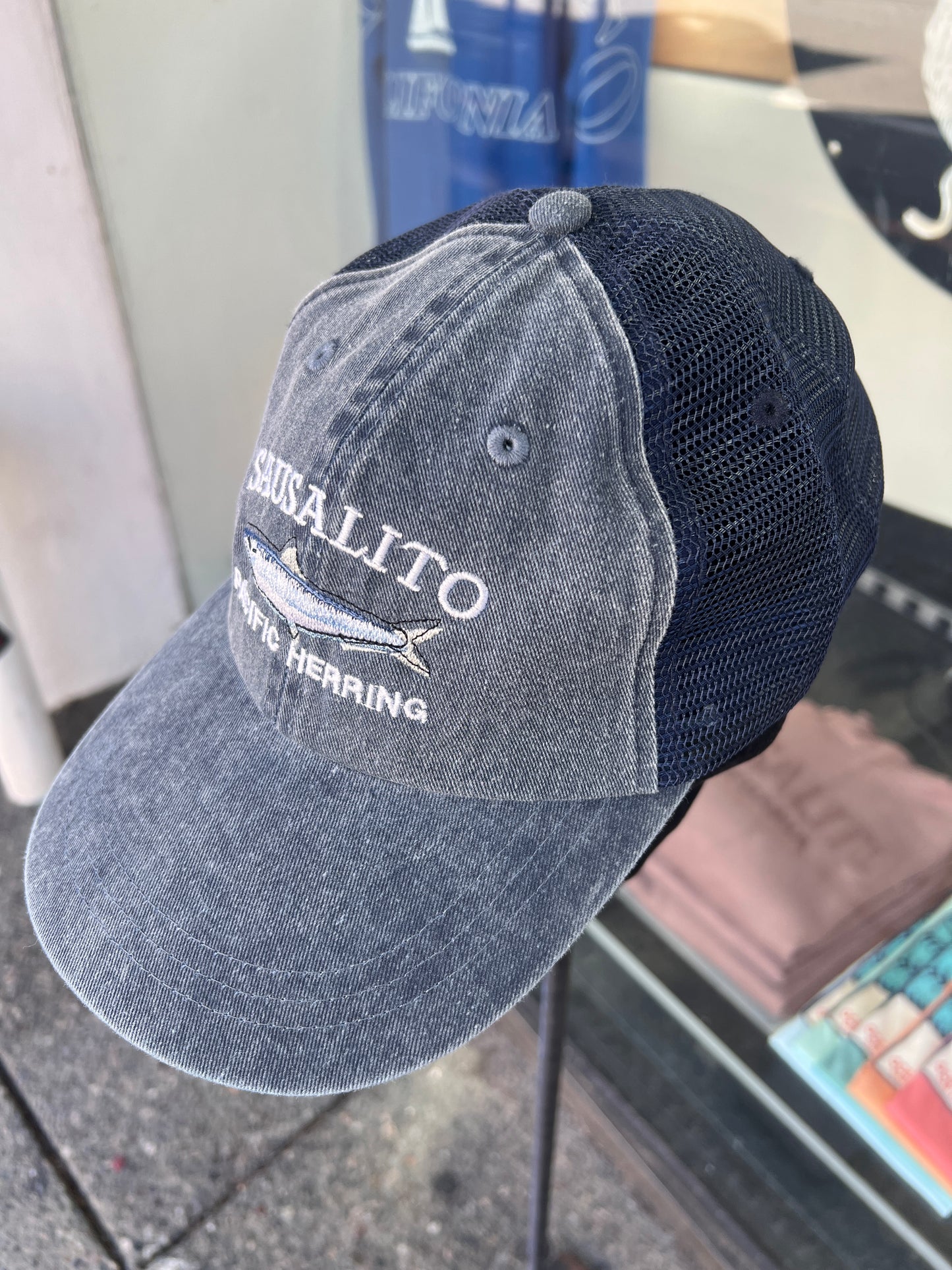 Denim washed cotton trucker hat featuring an embroidered white "SAUSALITO PACIFIC HERRING" design and a detailed silver-and-blue fish graphic.