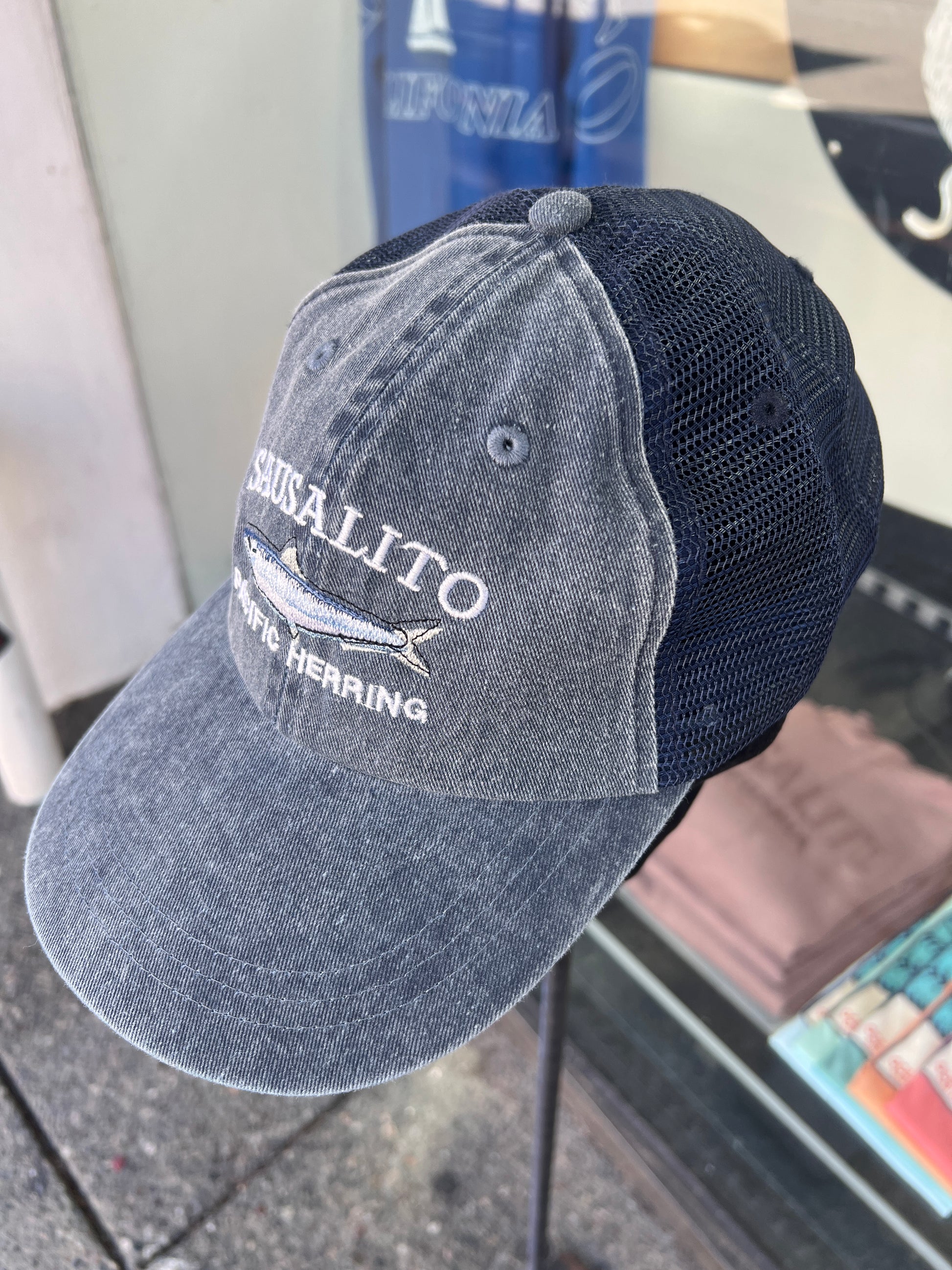 Denim washed cotton trucker hat featuring an embroidered white "SAUSALITO PACIFIC HERRING" design and a detailed silver-and-blue fish graphic.