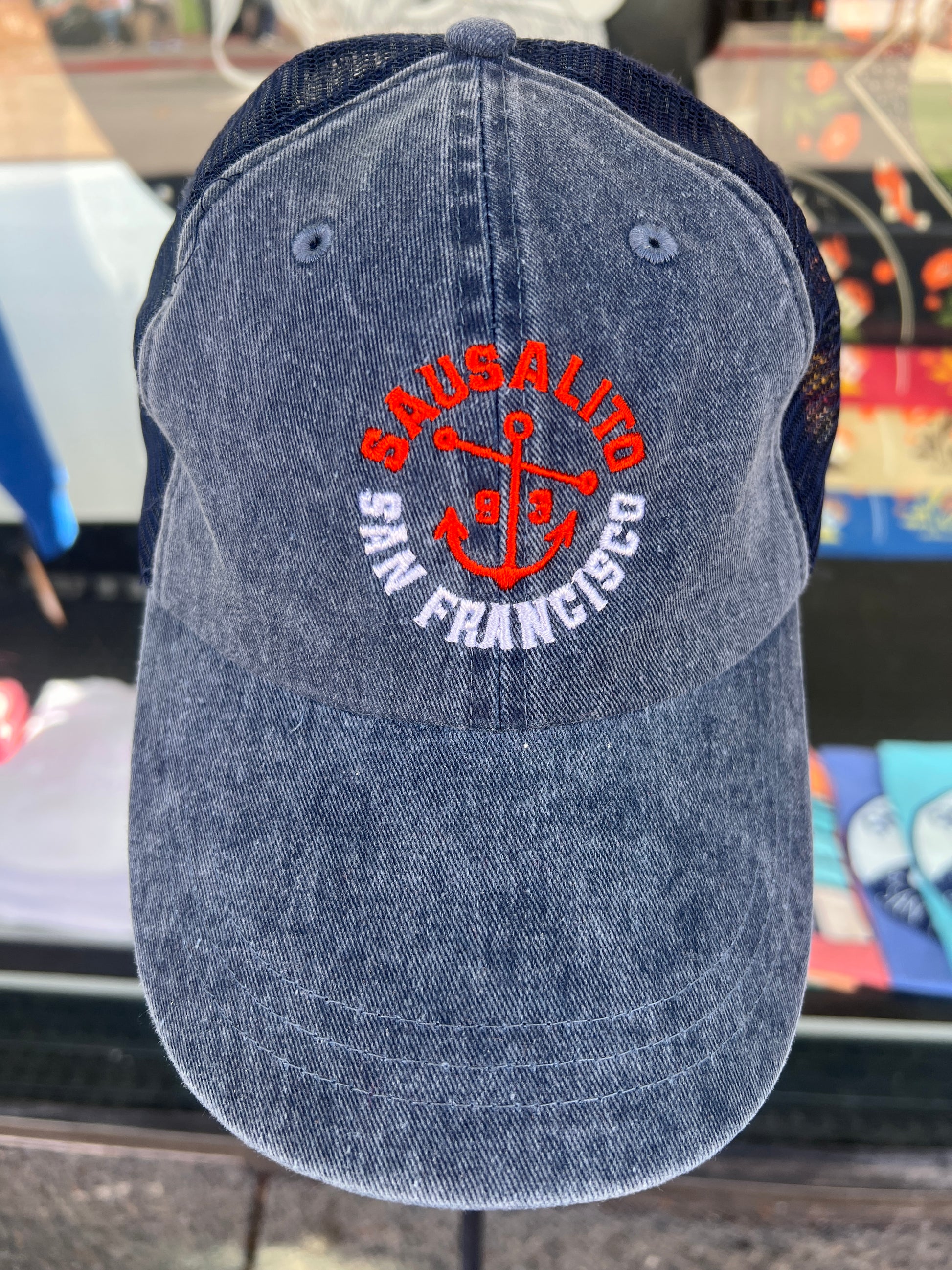 Blue washed denim trucker hat featuring an embroidered orange and white "SAUSALITO SAN FRANCISCO" anchor design with navy blue mesh back.