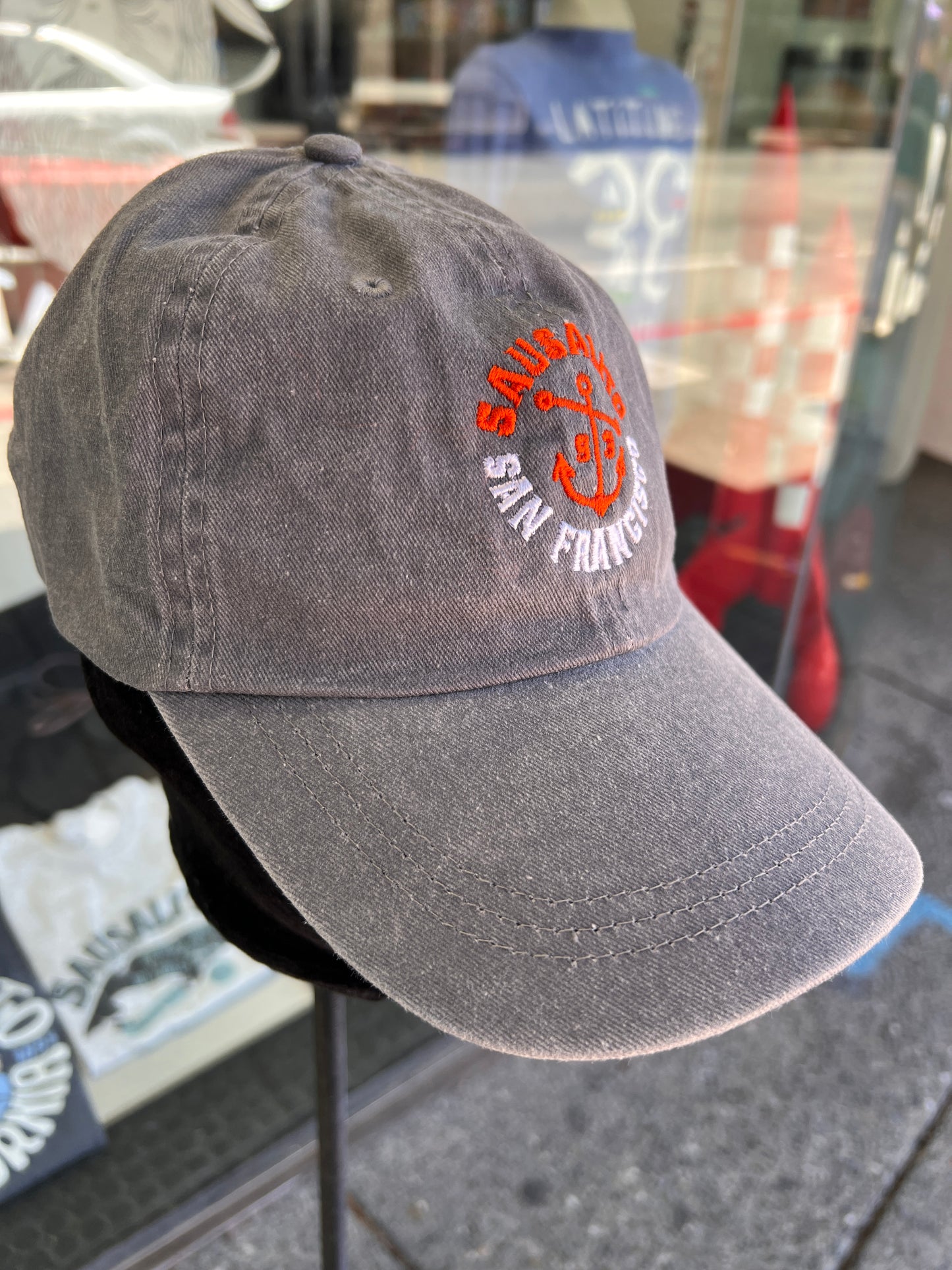 Side-angle view of a charcoal grey washed cotton baseball cap with an embroidered orange anchor and white text, displayed on a stand outside a Sausalito Ferry store window reflecting the street.