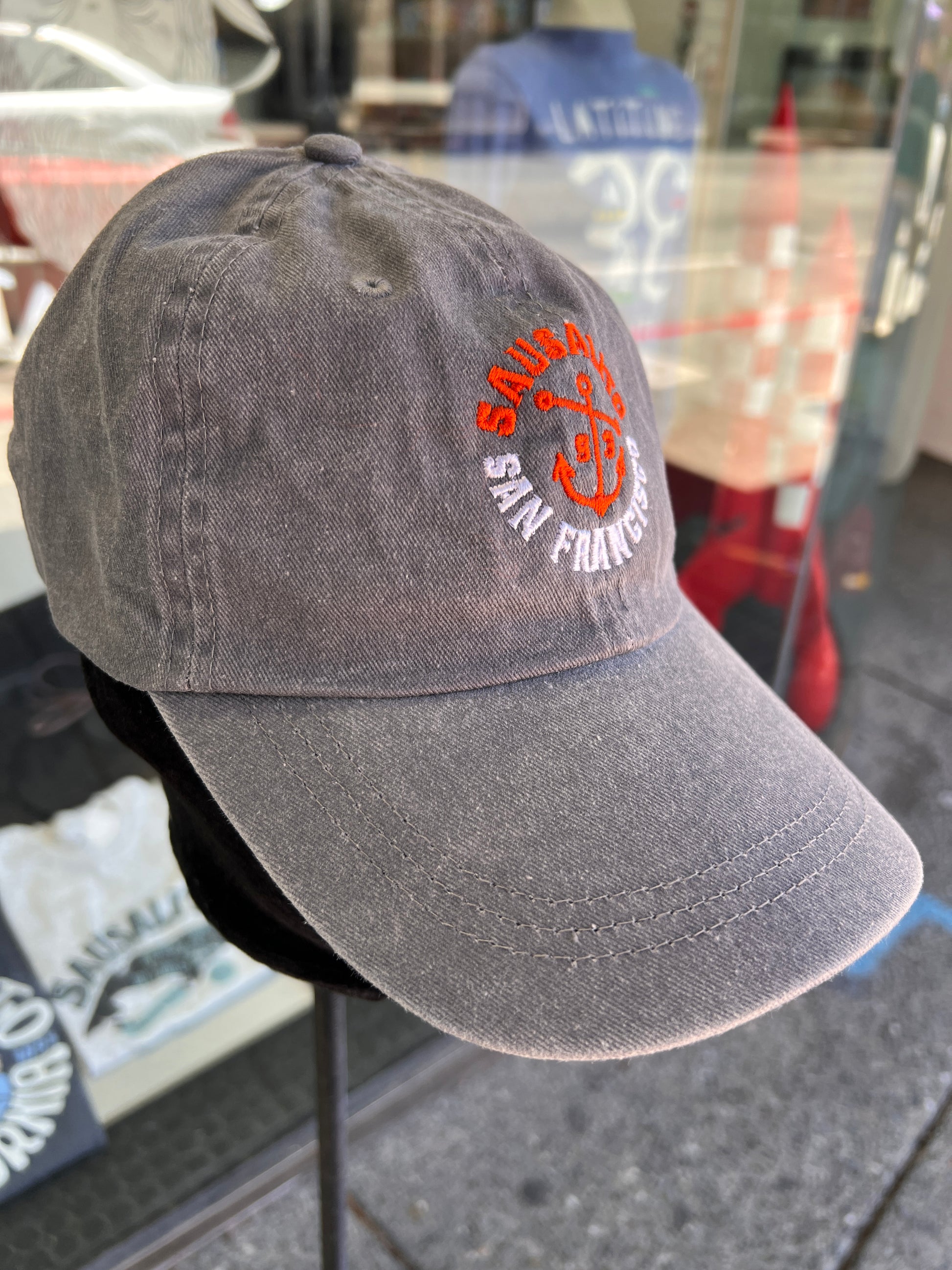 Side-angle view of a charcoal grey washed cotton baseball cap with an embroidered orange anchor and white text, displayed on a stand outside a Sausalito Ferry store window reflecting the street.
