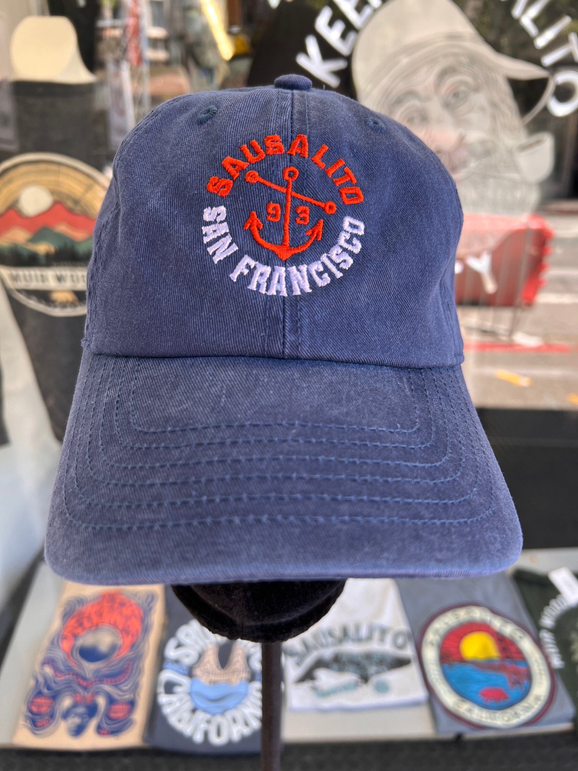 Front-facing view of a faded navy blue washed cotton cap featuring a custom embroidered orange anchor with white and orange typography reading Sausalito San Francisco 1893.