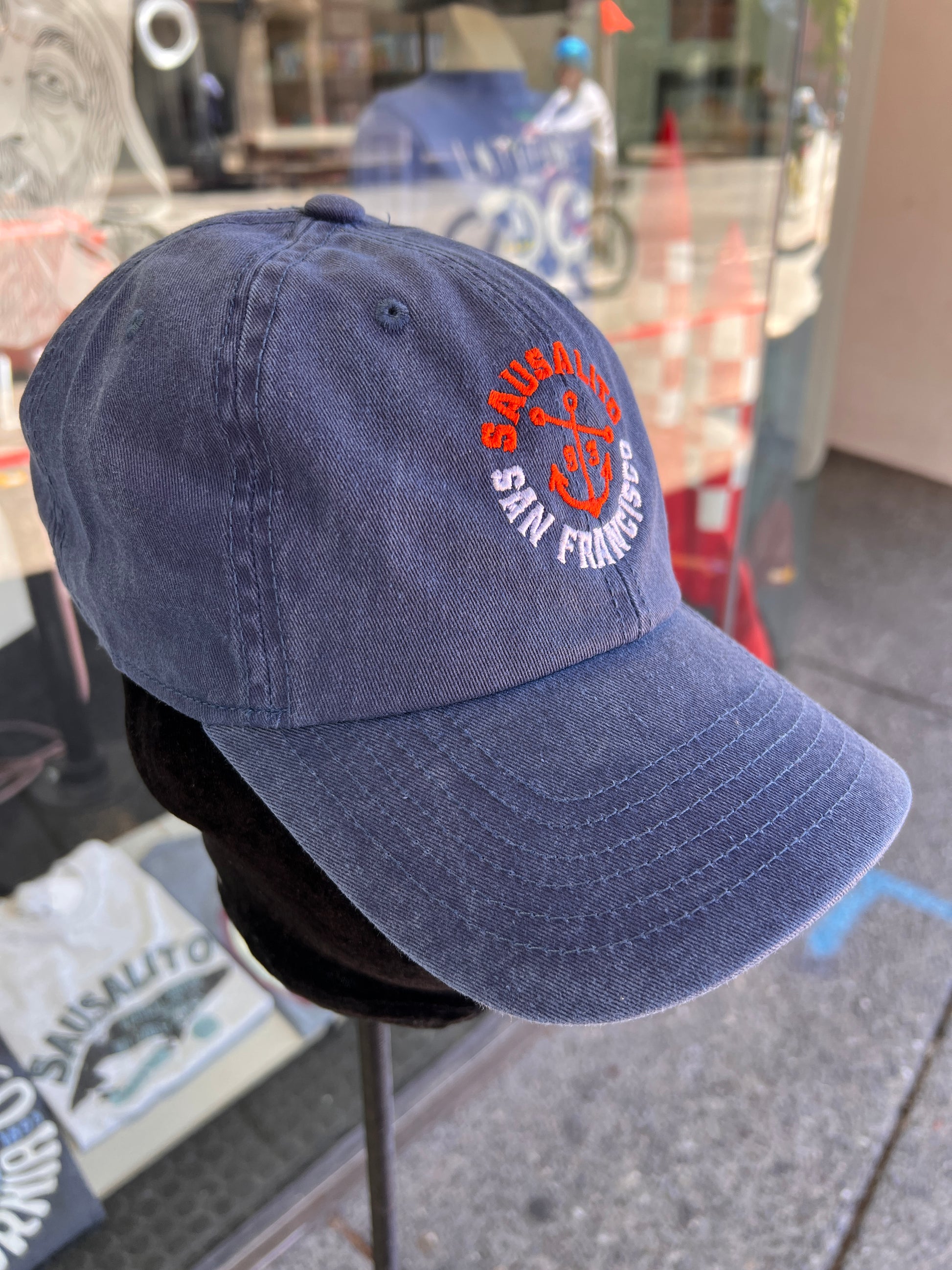 A close-up view of a navy blue washed cotton baseball cap featuring a custom embroidered orange anchor with 'Sausalito' in orange text above and 'San Francisco' in white text below, with '1893' as the incorporation year.