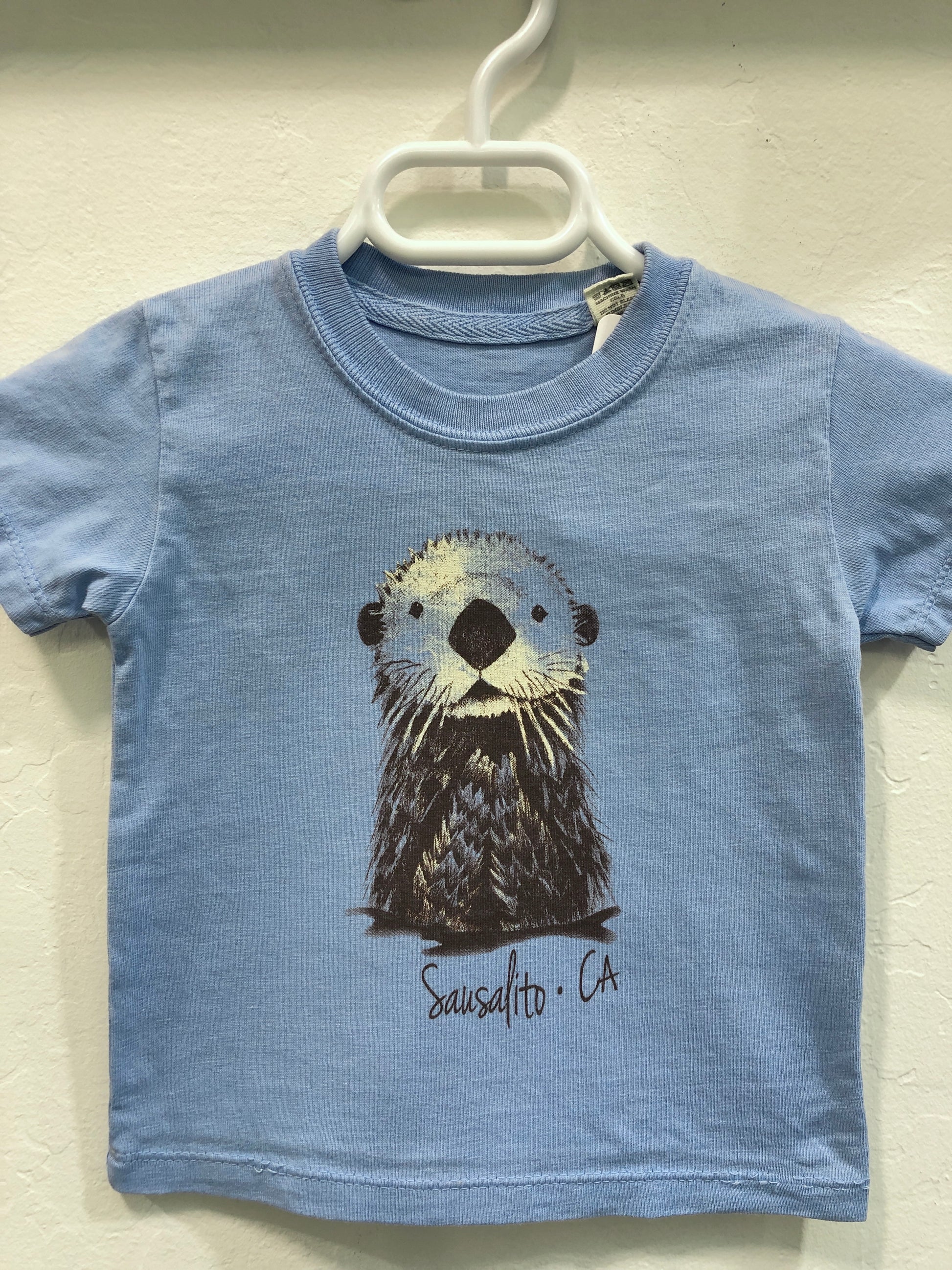 Blue infant t-shirt with cute sea otter illustration and 'Sausalito CA' text, hanging on white hanger