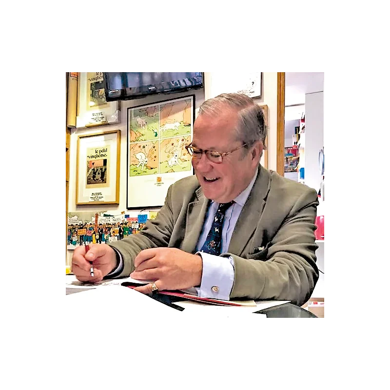 Tintin expert and author Michael Farr smiling while signing a copy of his book at a desk, with framed Tintin posters hanging on the wall behind him.