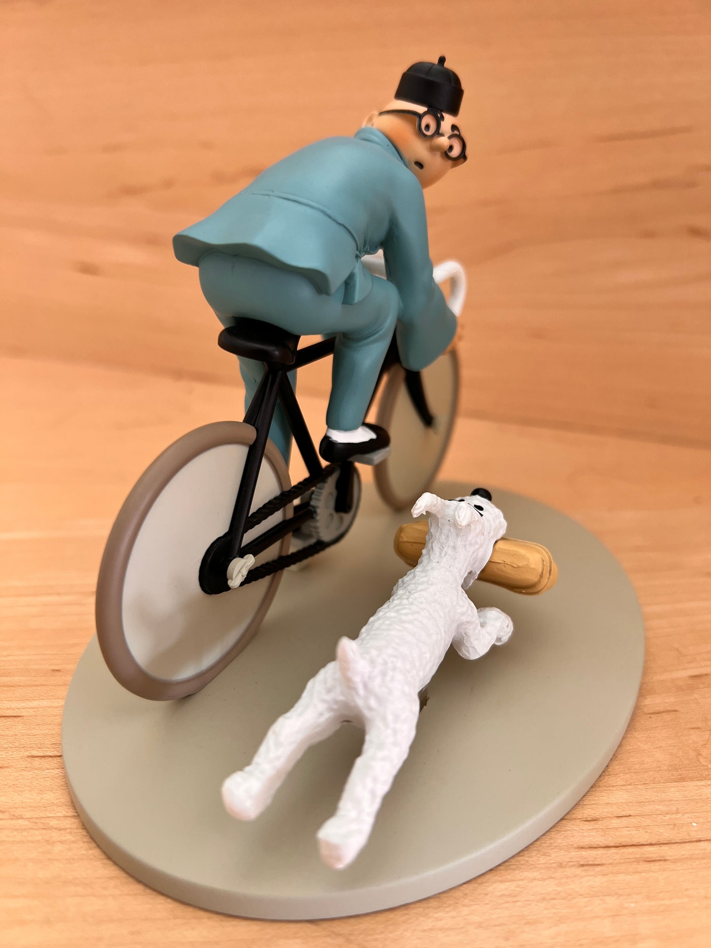 High-angle overhead view from behind the Tintin on a bicycle resin statuette, showing the back of Tintin's blue Chinese jacket as he pedals, with Snowy running close beside the rear wheel while carrying a tan package in his mouth on a light grey oval base.