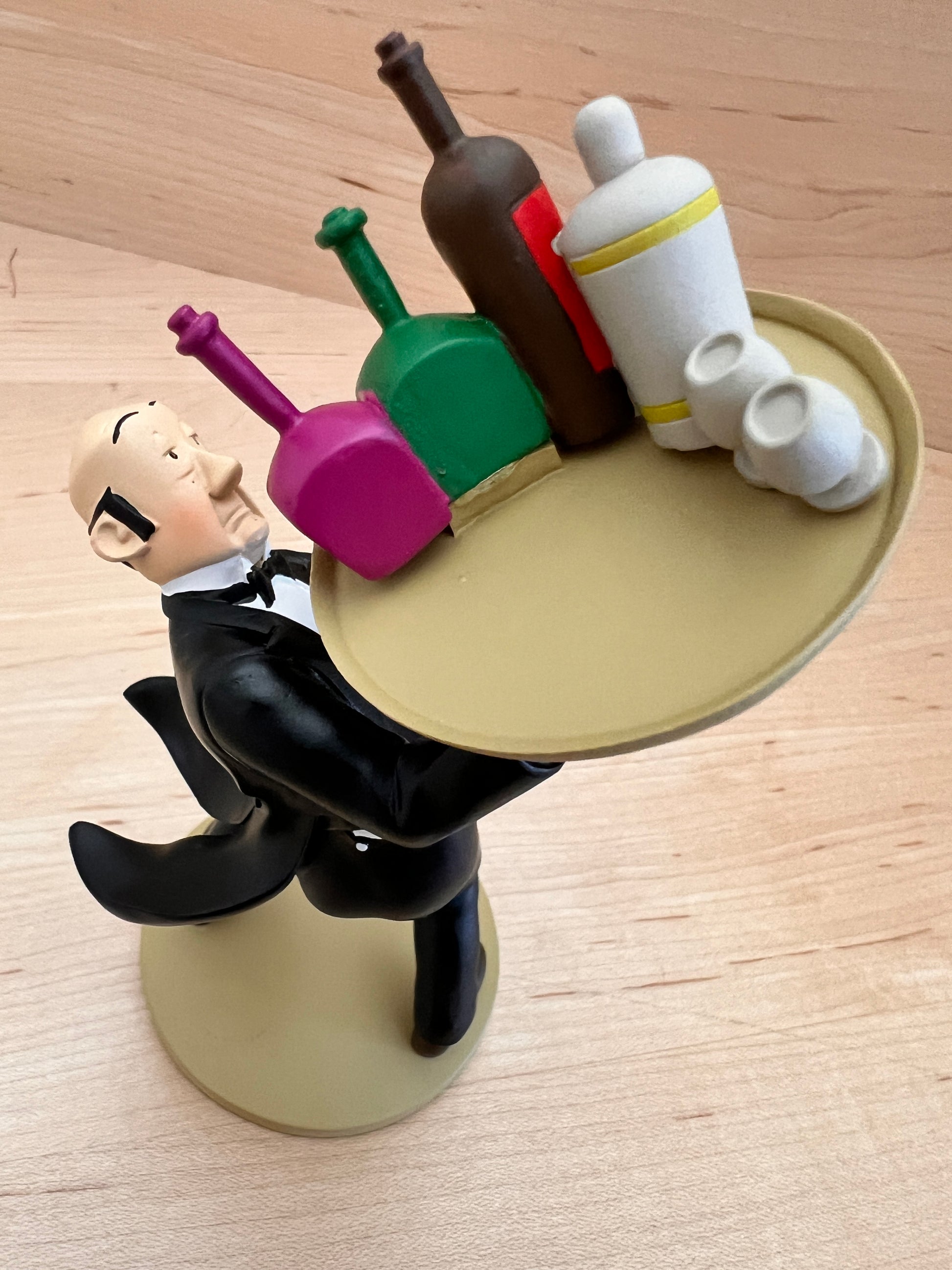 High-angle front view looking down at the Nestor resin figurine, detailing the butler's balding head, surprised expression, and black tuxedo coat as he balances a large round tan tray holding a white cocktail shaker, two small white cups, and three colorful liquor bottles.