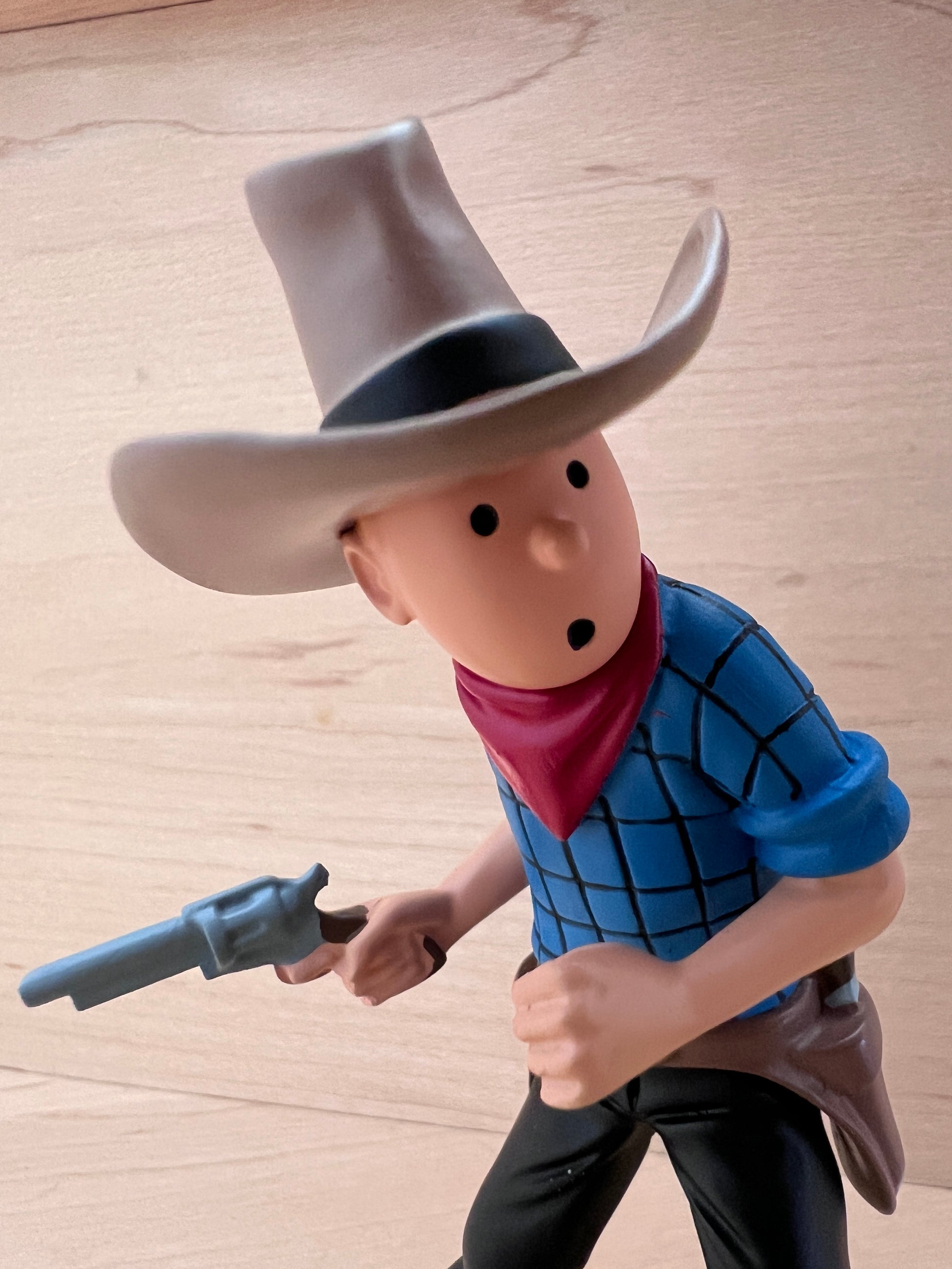 Close-up view of the Tintin in America polyresin figurine from the waist up, detailing Tintin's surprised facial expression, his wide-brimmed ten-gallon cowboy hat with a black band, a bright red bandana around his neck, and a blue-and-white checkered shirt while he holds a grey revolver pistol.