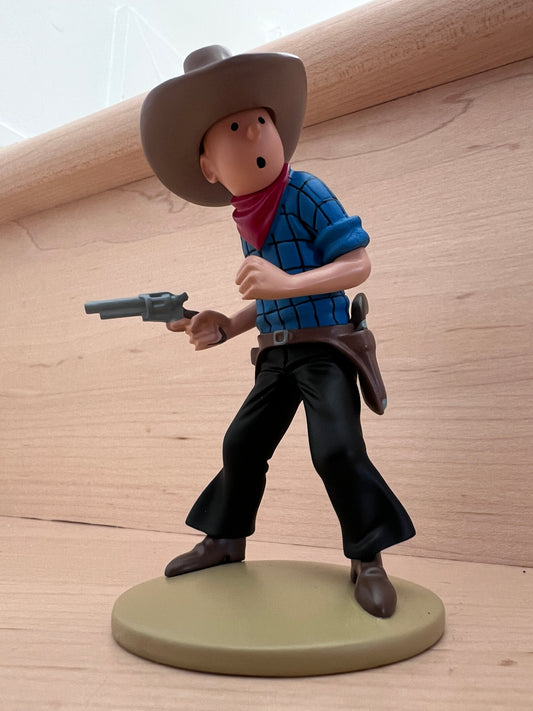 Tintin in America cowboy resin figure, 12 cm tall, wearing hat and hTintin in America hand-painted polyresin mini figure featuring Tintin dressed as a cowboy in a blue-and-white checkered shirt, black pants, a red bandana, and a ten-gallon hat, caught mid-action holding a six-shooter pistol on a round beige display base.