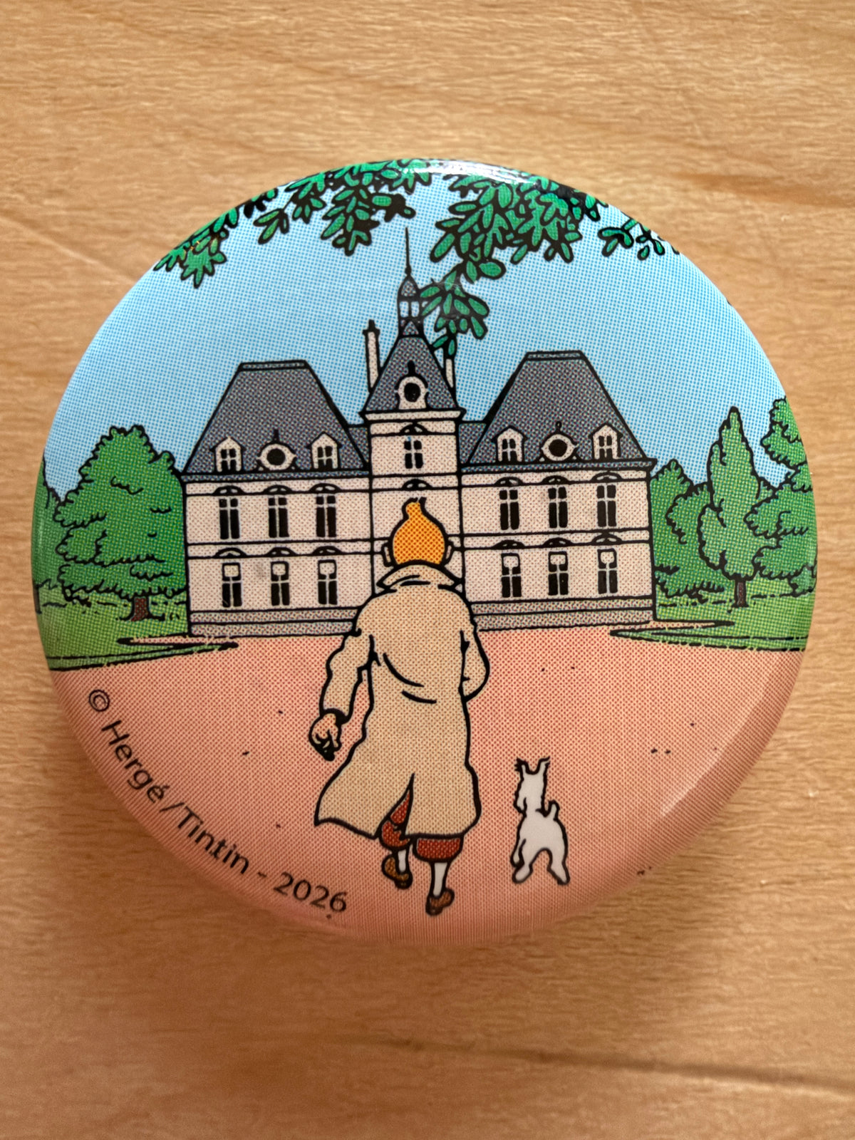 Close-up of a round 1-inch metal Tintin magnet featuring artwork of Tintin and Snowy walking toward Marlinspike Hall (Moulinsart) beneath a tree-lined pathway.