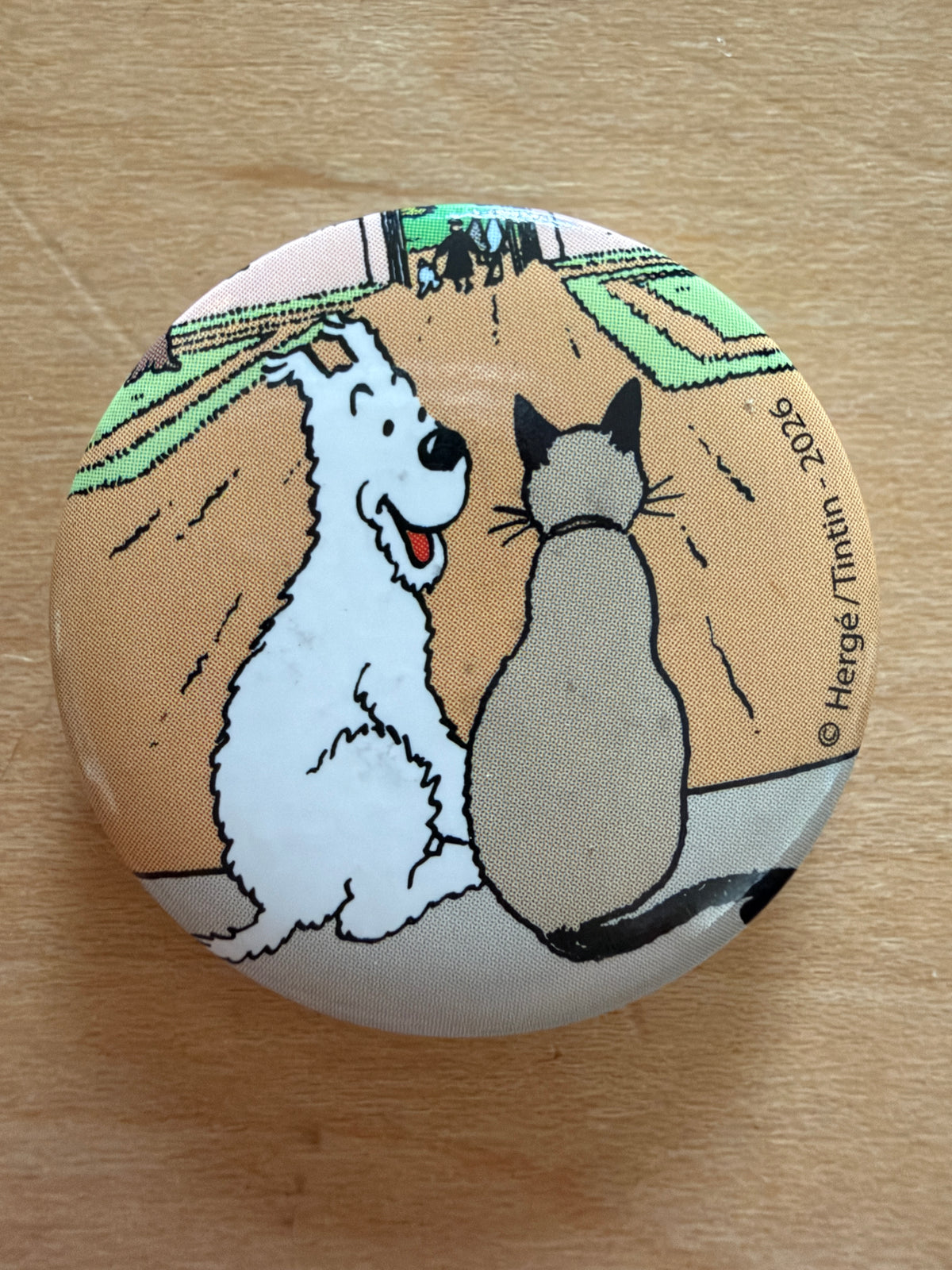 Close-up of a round 1-inch metal Tintin magnet featuring Snowy sitting next to a gray cat, with Tintin and another figure walking in the background.
