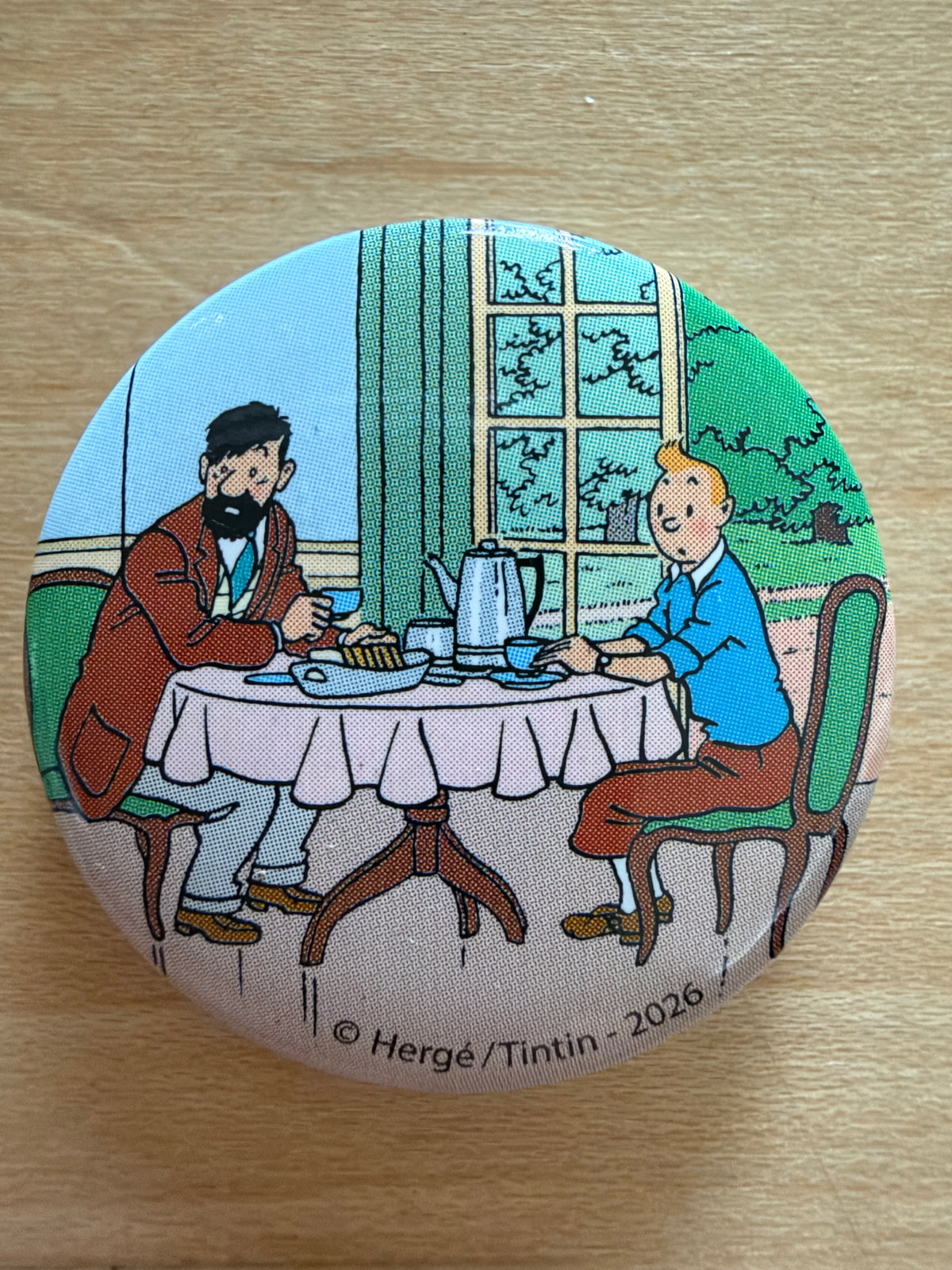 Close-up of a round 1-inch metal Tintin magnet featuring artwork from The Castafiore Emerald showing Captain Haddock and Tintin dining together at a table.