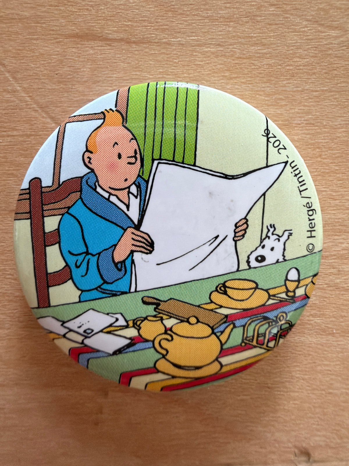 Close-up of a round 1-inch metal Tintin magnet featuring artwork of Tintin sitting in a blue robe reading a large newspaper at a breakfast table with Snowy peeking out beside him.