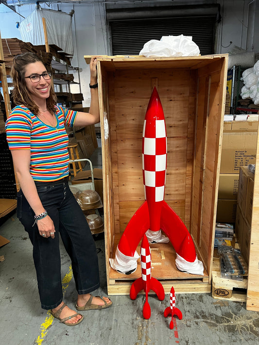 A person standing next to a large 150cm Tintin Moon Rocket model in a wooden shipping crate to demonstrate its scale, with smaller replica rockets on the floor in a warehouse setting.