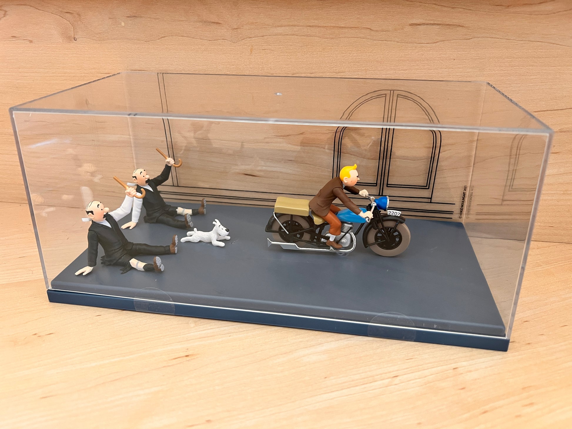 A Tintinimaginatio resin model from 'King Ottokar’s Sceptre' featuring Tintin on a motorcycle, housed inside a high-quality clear acrylic display case. The scene shows the detectives Thomson and Thompson on the ground behind the bike. The model is displayed on a light wooden surface.