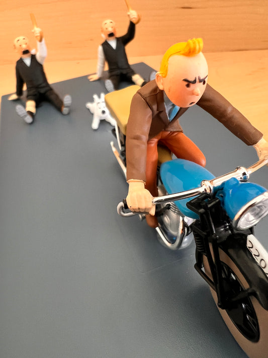 Close-up of a Tintinimaginatio resin model from 'King Ottokar’s Sceptre' featuring Tintin on a blue vintage motorcycle. In the background, Snowy and the detectives Thomson and Thompson are seen falling away as Tintin accelerates. High-detail collectible with hand-painted features.