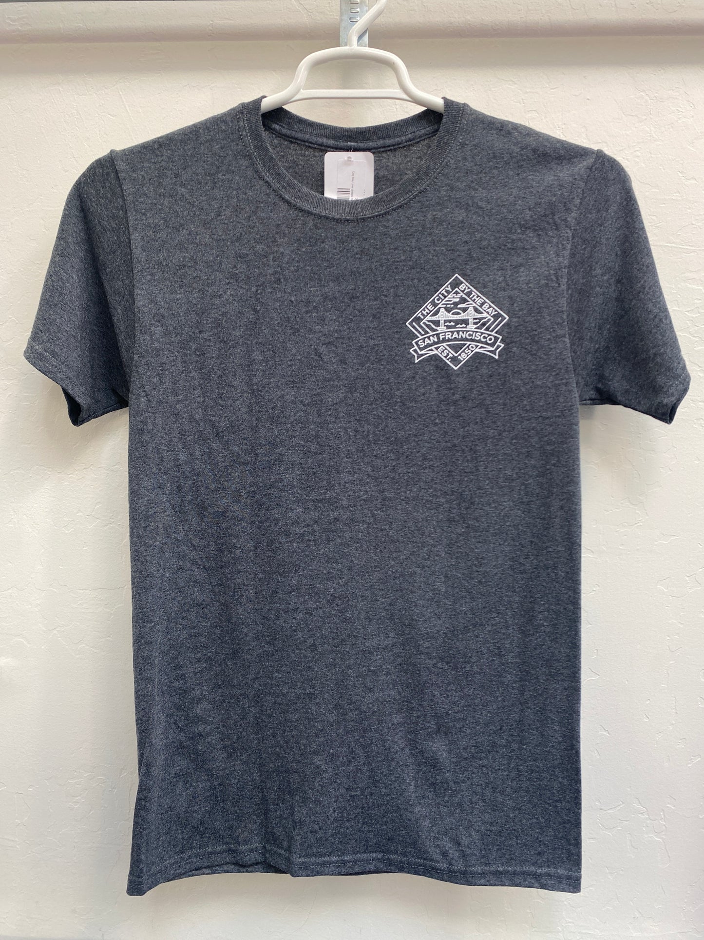 Front view of a charcoal gray heathered crewneck t-shirt hanging on a white plastic hanger against a light wall. The shirt features a clean, minimalist "City Bay Line" diamond-shaped emblem screen-printed in crisp white on the left chest. The graphic contains a stylized geometric outline of the Golden Gate Bridge under a setting sun over the bay, framed by the text "THE CITY BY THE BAY" on top and "SAN FRANCISCO" across the center.
