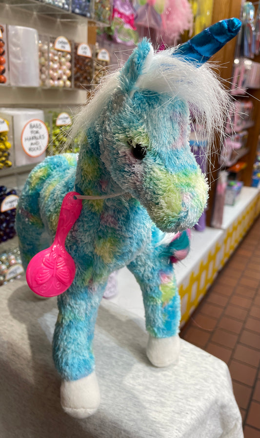 Colorful plush unicorn toy with blue coat, shimmery horn, and long mane, holding pink brush in store display