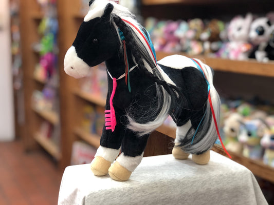 Plush black and white pinto horse toy with colorful ribbons in mane and tail, featuring a pink brush accessory