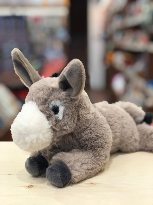 Soft gray floppy donkey plush toy with white muzzle and dark hooves, sitting on wooden surface in toy store