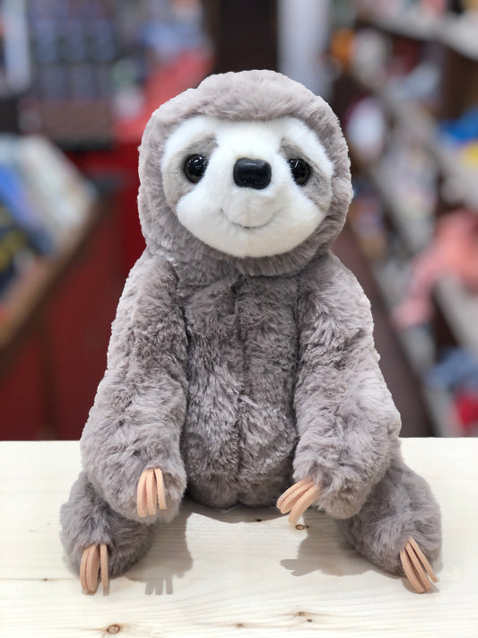 Cuddly gray sloth plush toy with soft fur, smiling face, and felt claws sitting on white surface in toy store