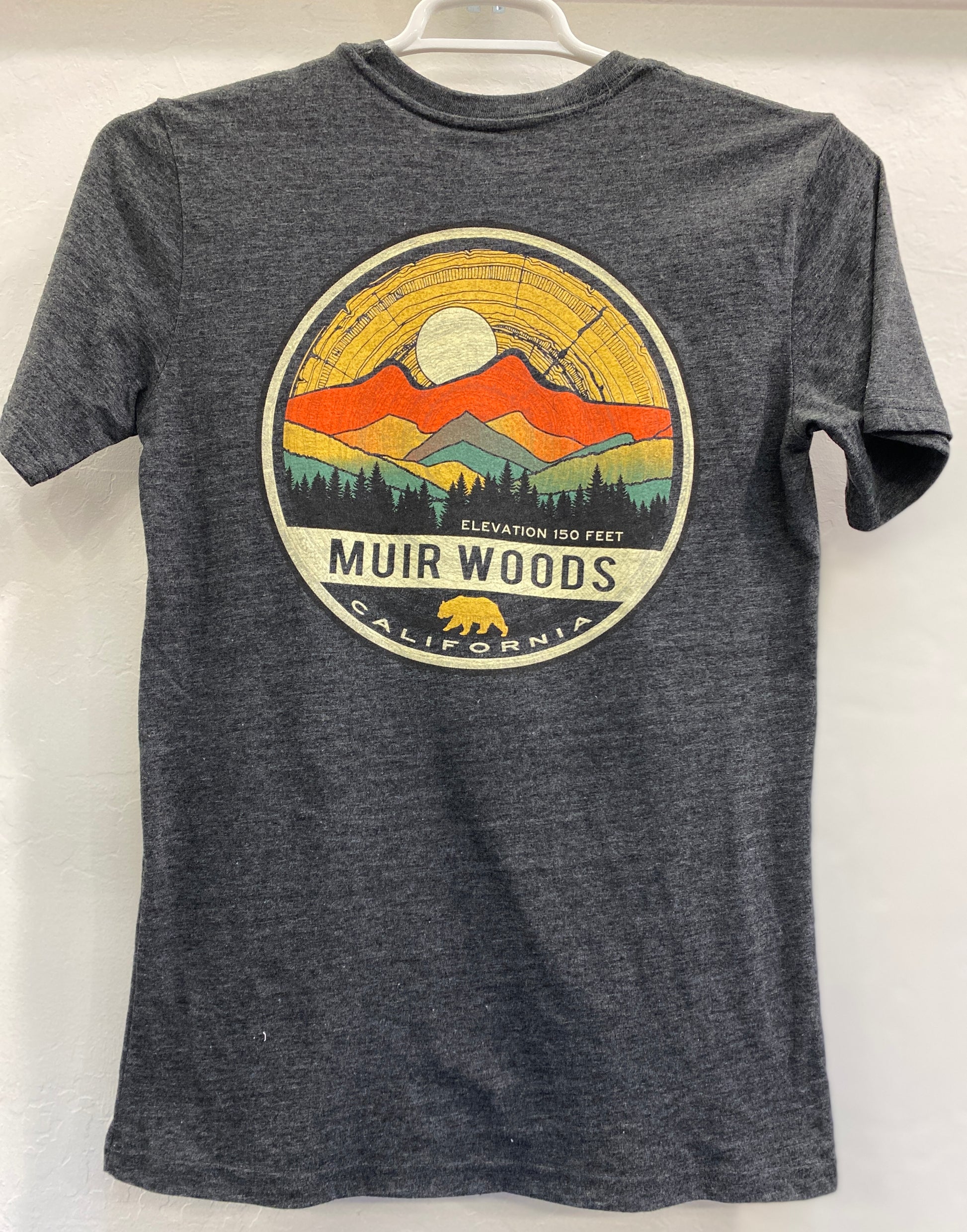 Charcoal gray heather unisex t-shirt featuring a circular graphic emblem on the front with a yellow full moon, orange and yellow mountains, a green silhouette of a redwood forest, and a gold California bear logo at the bottom with the text "Muir Woods California, Elevation 150 Feet".