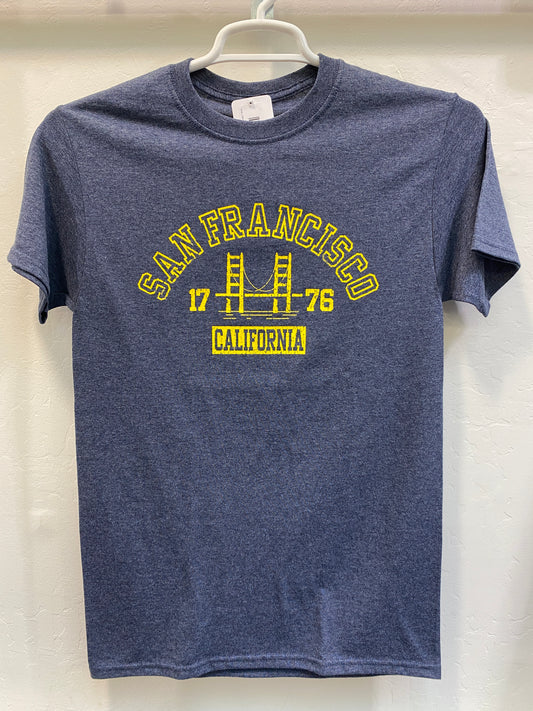 Front view of a navy blue heathered crewneck t-shirt hanging on a white plastic hanger against a light, textured wall. The shirt features a vintage collegiate-style graphic screen-printed on the chest in vibrant golden yellow. The design features the words "SAN FRANCISCO" arched over a minimalist, architectural silhouette of the Golden Gate Bridge. The year "17" and "76" are split on either side of the bridge towers, and the word "CALIFORNIA" is enclosed in a solid yellow rectangular block directly undernea