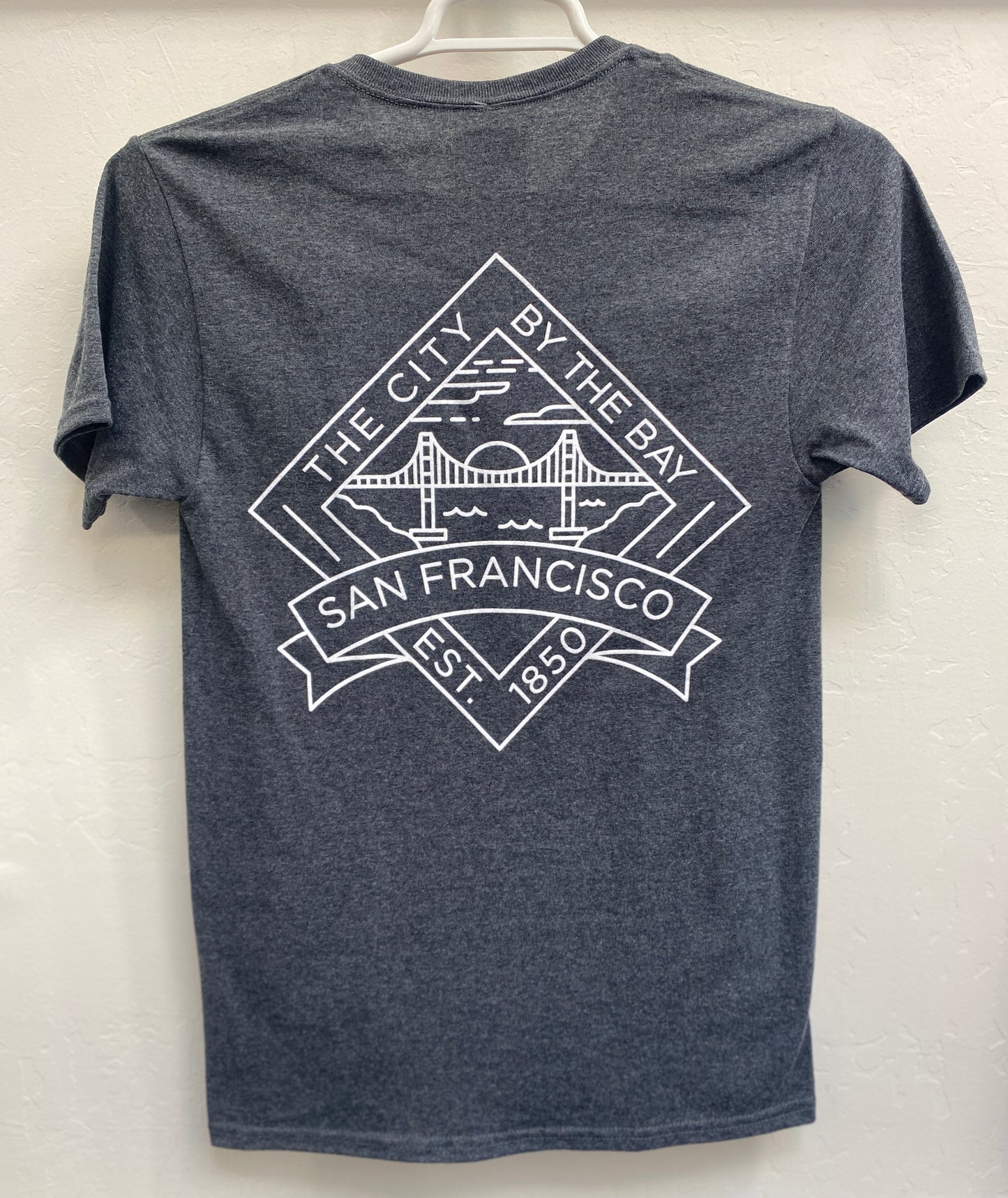 Back view of a charcoal gray heathered crewneck t-shirt hanging on a white plastic hanger. The shirt features a large, crisp white geometric graphic screen-printed across the upper back. The design is a diamond-shaped crest containing a minimalist outline of the Golden Gate Bridge under a setting sun over the bay, framed by the arched text "THE CITY BY THE BAY" on top, "SAN FRANCISCO" on a bold ribbon banners across the center, and "EST. 1850" at the bottom point.
