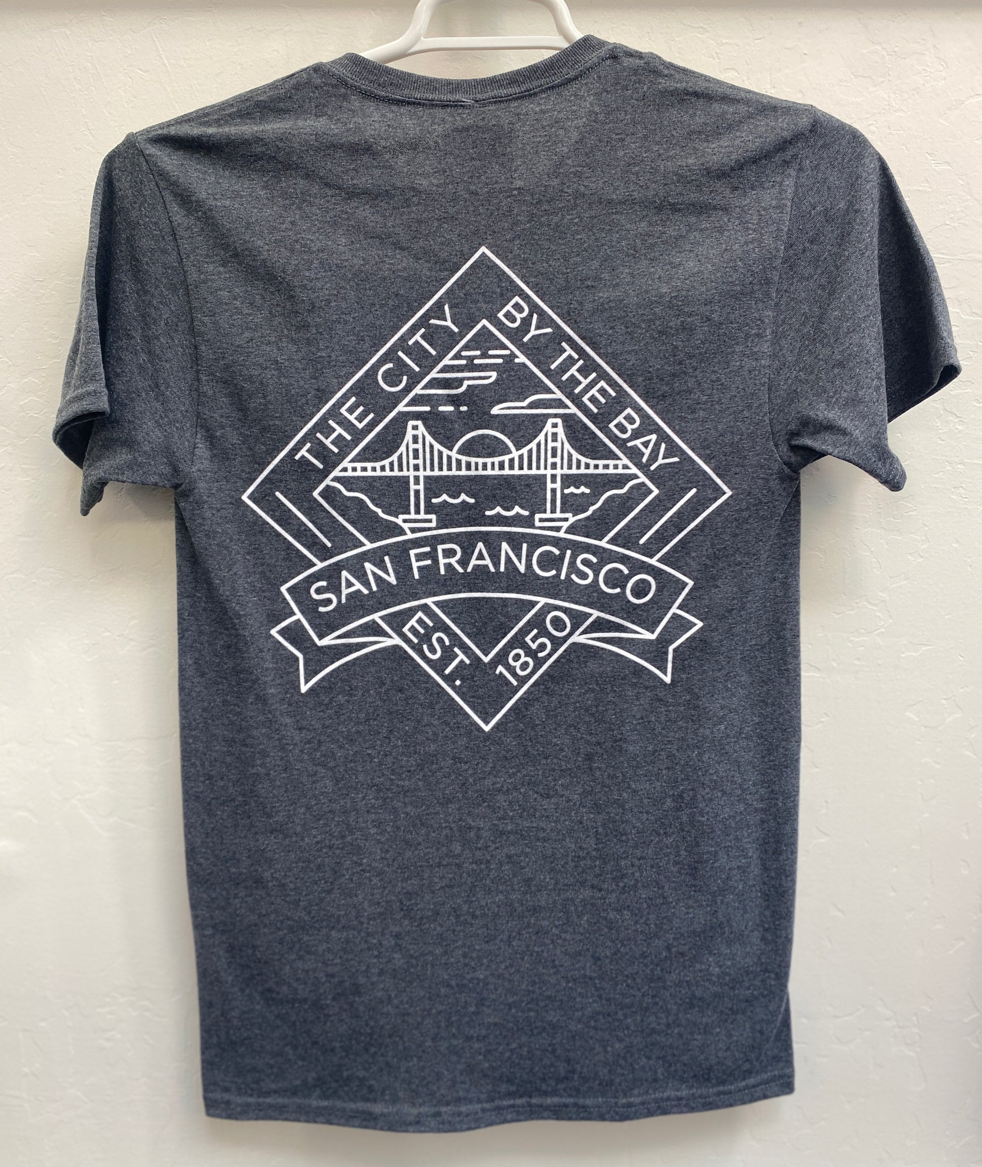 Back view of a charcoal gray heathered crewneck t-shirt hanging on a white plastic hanger. The shirt features a large, crisp white geometric graphic screen-printed across the upper back. The design is a diamond-shaped crest containing a minimalist outline of the Golden Gate Bridge under a setting sun over the bay, framed by the arched text "THE CITY BY THE BAY" on top, "SAN FRANCISCO" on a bold ribbon banners across the center, and "EST. 1850" at the bottom point.