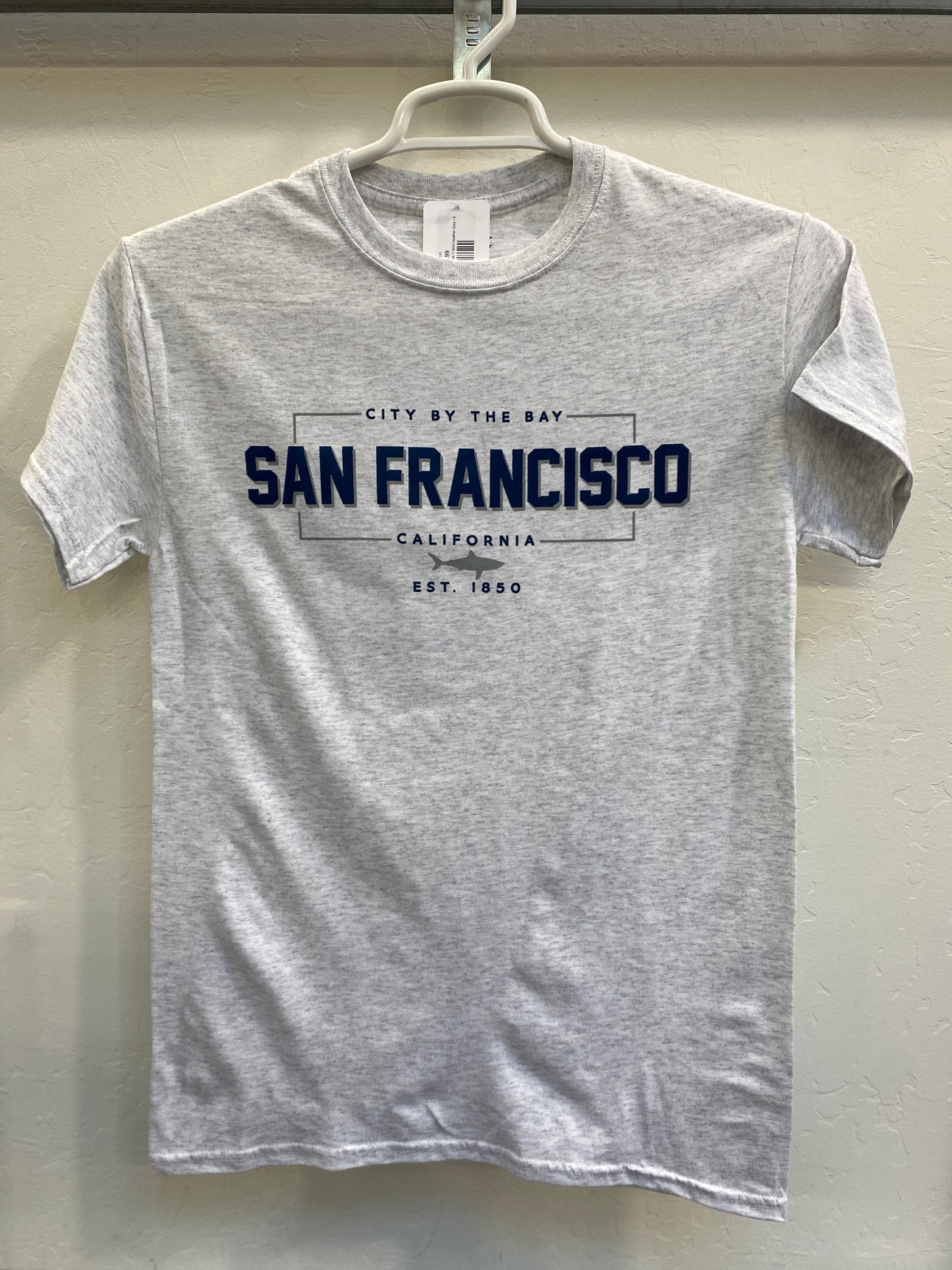 Front view of a heather gray crewneck t-shirt hanging on a white plastic hanger against a light-colored wall. The shirt features a dark blue and gray athletic-style graphic on the chest. The design features a crisp rectangular border enclosing the bold text "SAN FRANCISCO", with "CITY BY THE BAY" arched cleanly above it and "CALIFORNIA" written below. A small, detailed silhouette of a great white shark is centered directly beneath the city name, anchored by the vintage font text "EST. 1850".