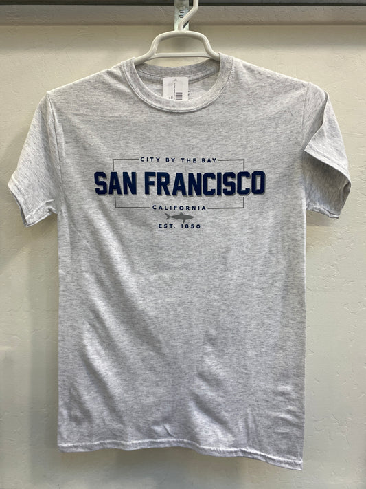 Front view of a heather gray crewneck t-shirt hanging on a white plastic hanger against a light-colored wall. The shirt features a dark blue and gray athletic-style graphic on the chest. The design features a crisp rectangular border enclosing the bold text "SAN FRANCISCO", with "CITY BY THE BAY" arched cleanly above it and "CALIFORNIA" written below. A small, detailed silhouette of a great white shark is centered directly beneath the city name, anchored by the vintage font text "EST. 1850".