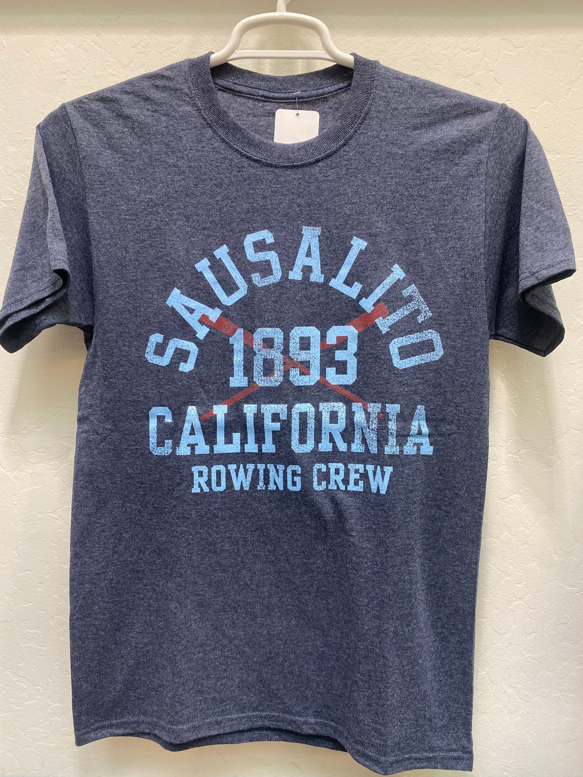 Sausalito 1893 California Rowing Crew t-shirt in navy blue with light blue text and crossed red oarsFront view of a navy blue heathered crewneck t-shirt hanging on a white plastic hanger against a white wall. The shirt features a large, retro athletic graphic screen-printed on the chest with the text "SAUSALITO" arched over "CALIFORNIA" and "ROWING CREW" in light blue. Centered in the middle is the year "1893" stamped over two bold, crossed oars in a vibrant crimson red.