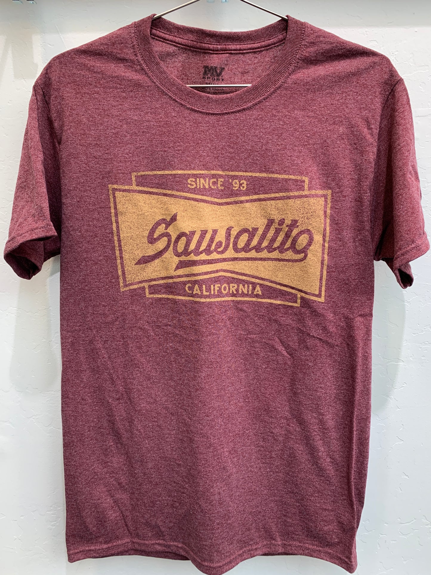 Front view of a maroon heathered crewneck t-shirt hanging on a wire hanger against a white textured wall. The shirt features a large, gold screen-printed graphic on the chest shaped like a classic bow tie or banner frame. Inside the border, stylized vintage-style script reads "Sausalito", anchored by "SINCE '93" along the top edge and "CALIFORNIA" centered along the bottom.