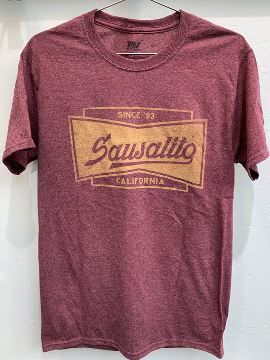 Front view of a maroon heathered crewneck t-shirt hanging on a wire hanger against a white textured wall. The shirt features a large, gold screen-printed graphic on the chest shaped like a classic bow tie or banner frame. Inside the border, stylized vintage-style script reads "Sausalito", anchored by "SINCE '93" along the top edge and "CALIFORNIA" centered along the bottom.