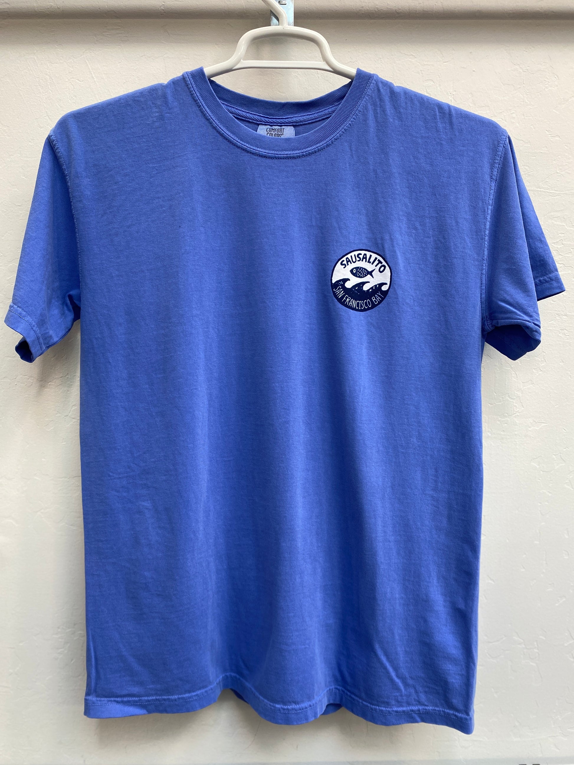 Close-up of a blue cotton t-shirt featuring a small, circular white and dark blue breast logo with a stylized fish leaping over waves and the text "Sausalito San Francisco Bay".