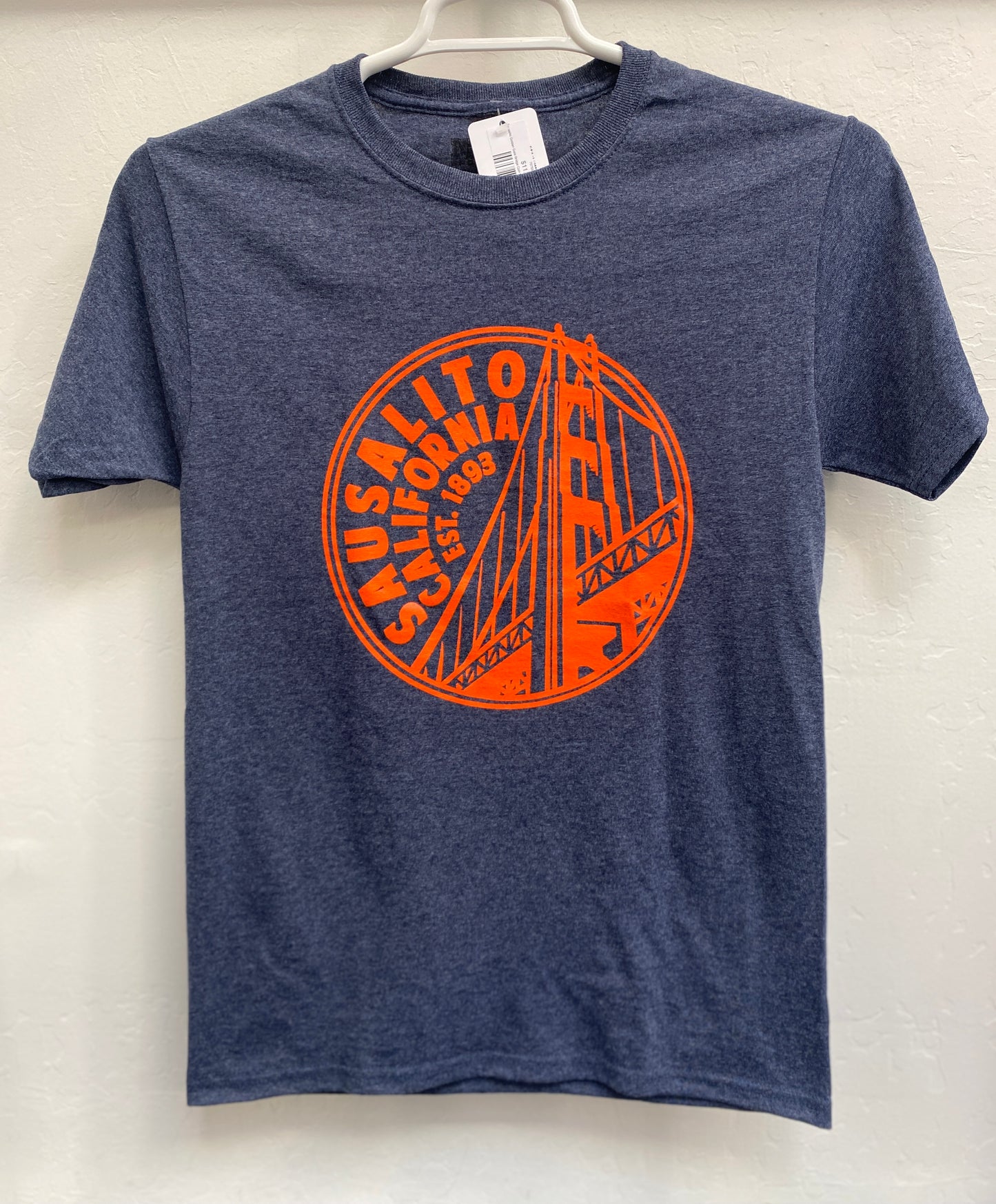 Front view of a navy blue heathered crewneck t-shirt hanging on a w hanger against a textured wall. The shirt features a large, vibrant Golden Gate Orange screen-printed circular graphic on the chest. The design showcases a stylized silhouette of the Golden Gate Bridge's north span tower, framed by bold curved text reading "SAUSALITO CALIFORNIA" and anchored by the incorporation year "EST. 1893".