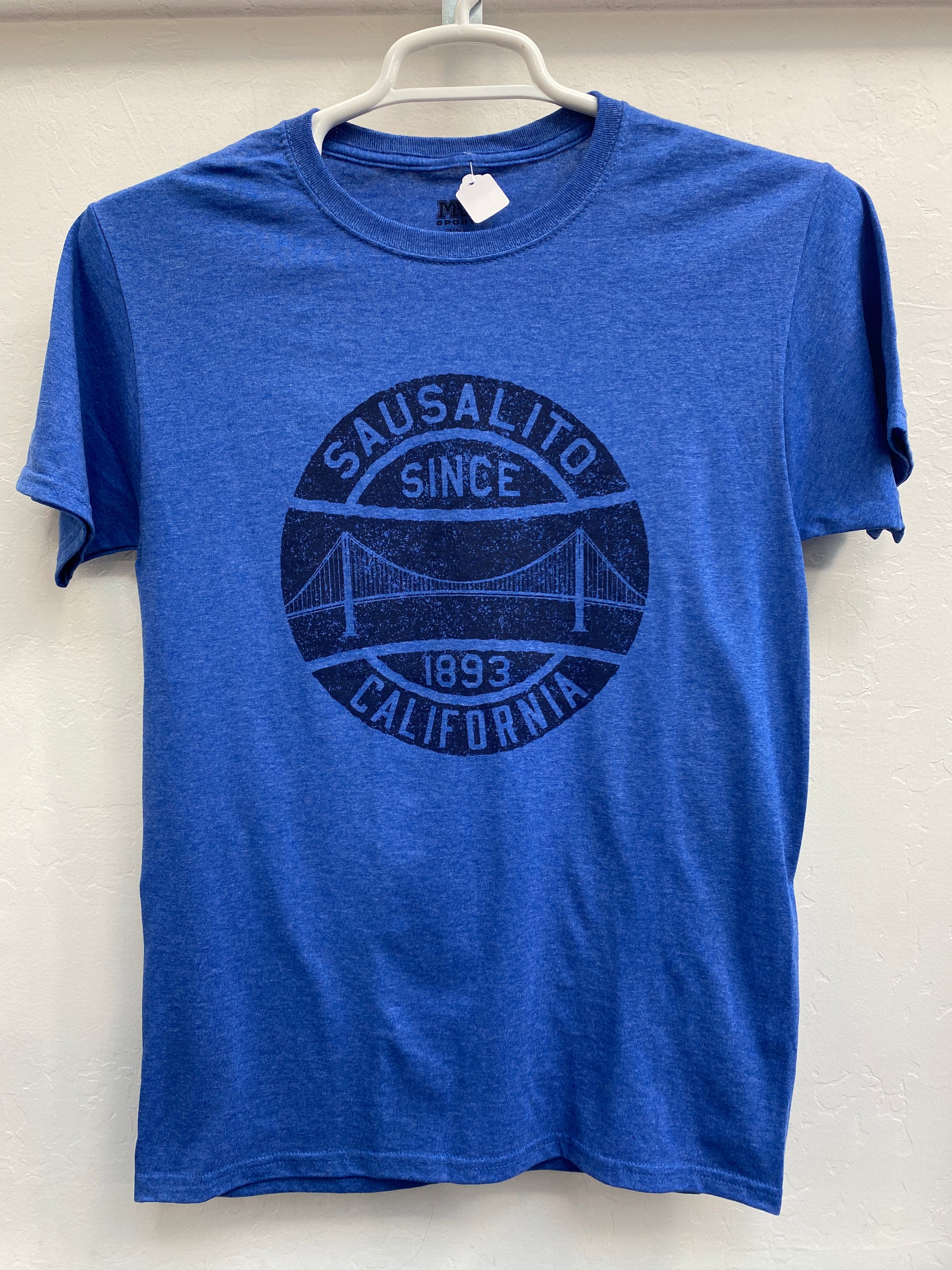 Front view of a heather blue crewneck t-shirt hanging on a white plastic hanger against a light, neutral background. The shirt features a large, dark blue circular graphic printed on the chest. The design features a stylized, clean geometric silhouette of the Golden Gate Bridge under an open sky, framed inside a dual-ring border with the bold text "SAUSALITO" arched along the top, "SINCE 1893" running across the lower middle, and "CALIFORNIA" anchoring the bottom curve.