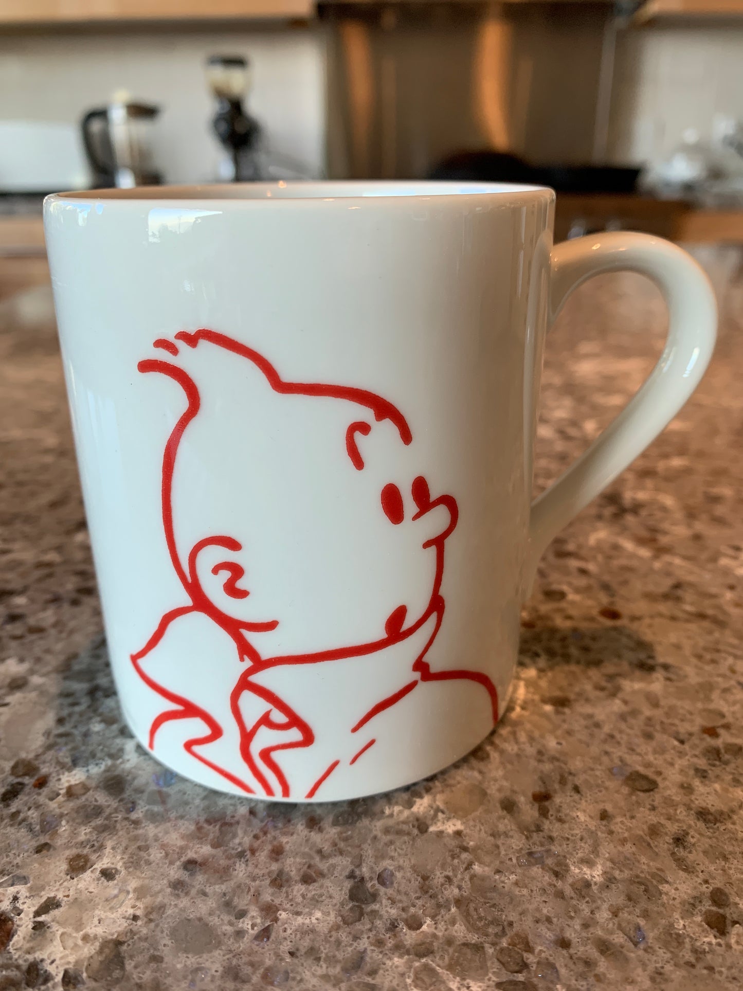 Close-up of the white porcelain Surprised Tintin mug on a speckled brown granite countertop, featuring a bold, minimalist red line-art illustration of Tintin's iconic profile with a wide-eyed, startled expression.